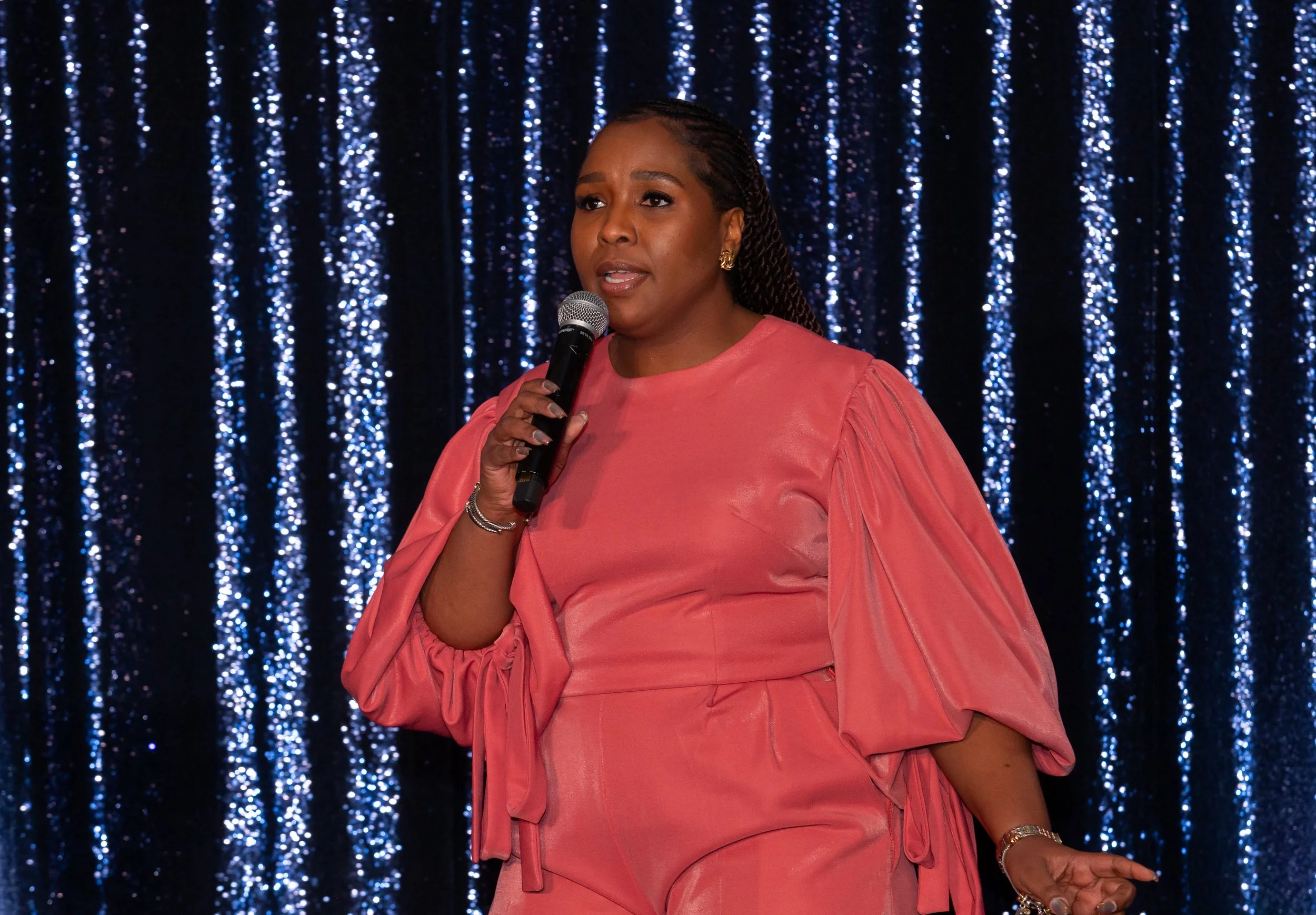A woman in a pink dress holding a microphone, speaking on stage with a dark, sparkly blue curtain background.