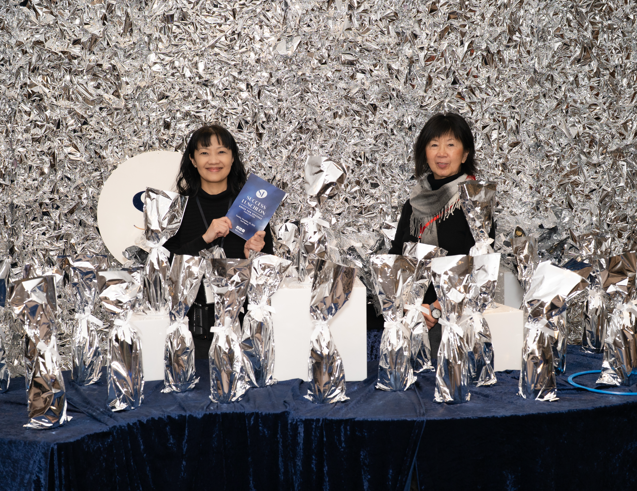 Two women standing behind a table with numerous silver-wrapped objects, against a backdrop of crumpled silver foil. One woman holds a blue paper, smiling, and the other woman faces slightly to the side.