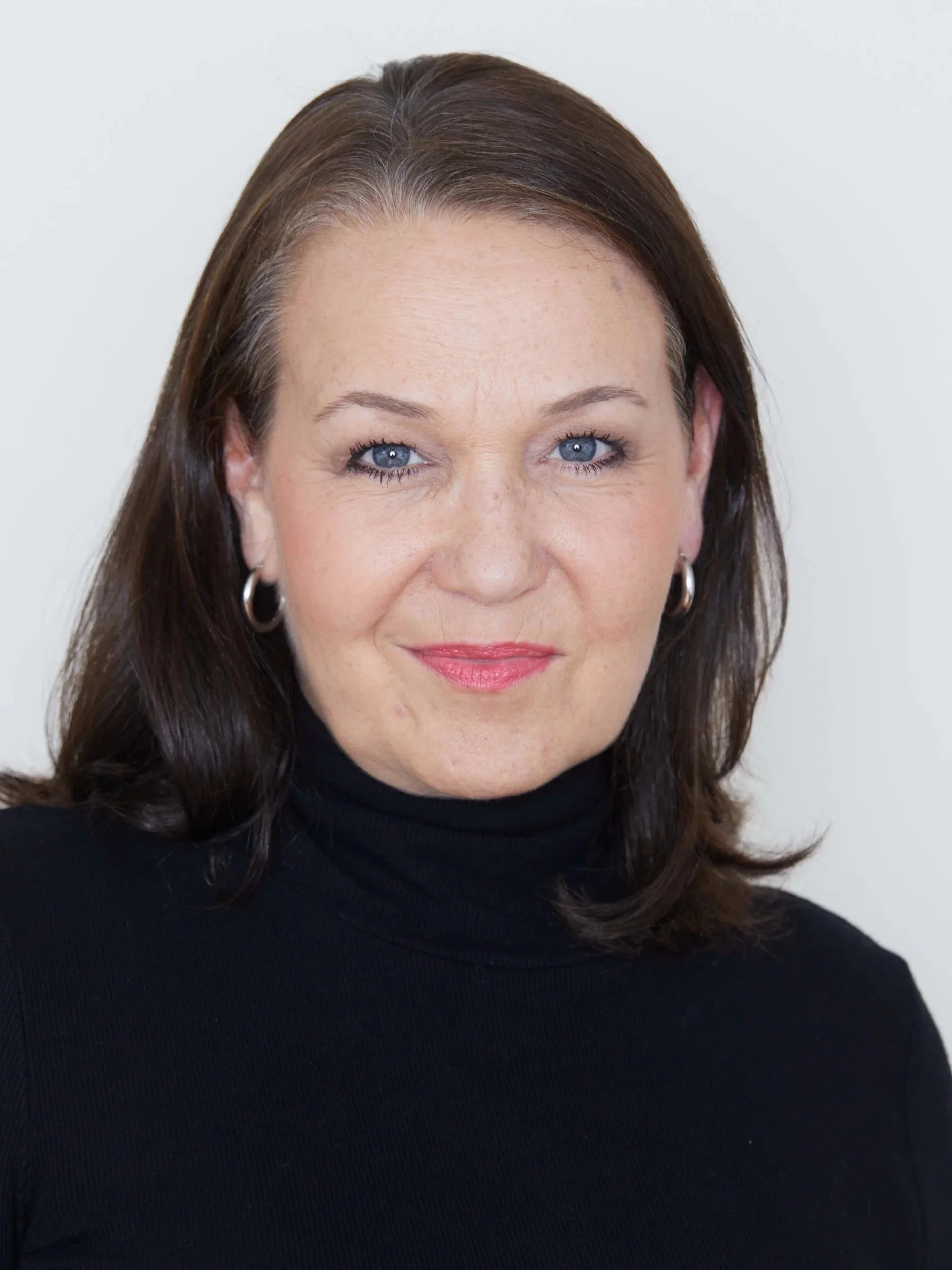 A woman with shoulder-length brown hair, blue eyes, and hoop earrings wearing a black turtleneck, smiling slightly against a light background.