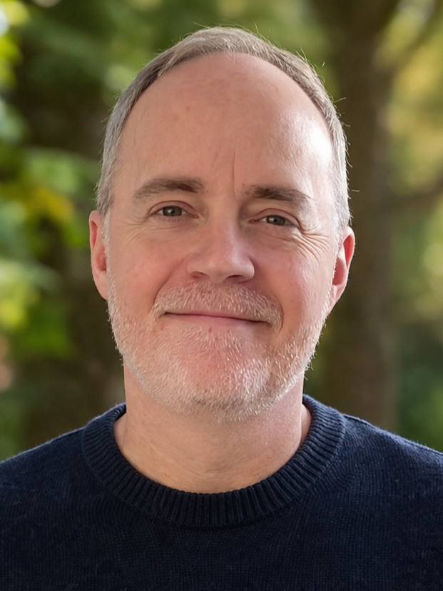 A middle-aged man outdoors with a gentle smile, wearing a dark sweater, with a blurred natural background of green trees and soft sunlight.