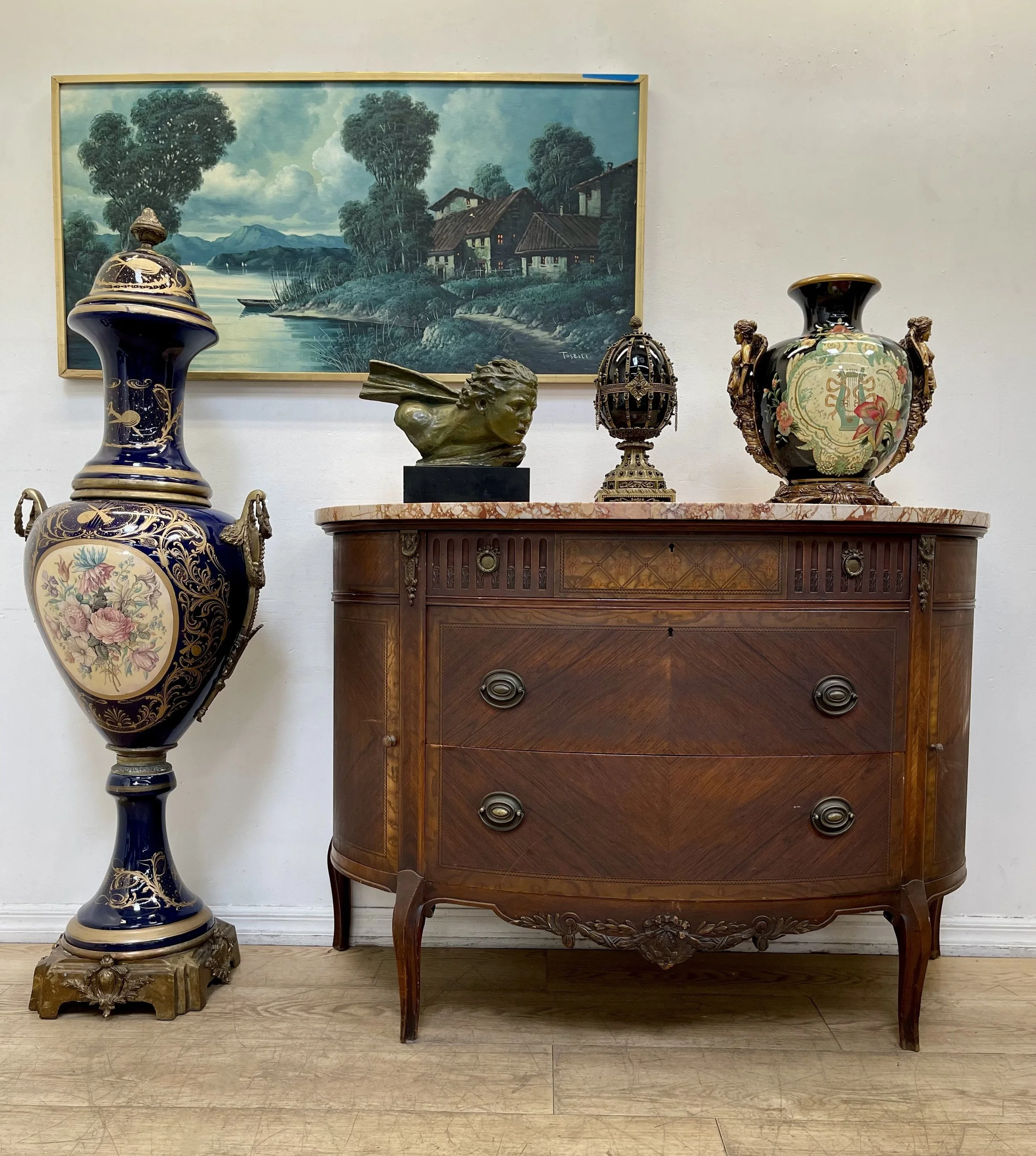 Mid-century modern room with twin orange leather chairs, a wooden sideboard decorated with vases, sculptures, and decorative objects. A colorful floral area rug is on the floor, and a vintage floor lamp stands on the left. A folding screen with botanical designs and a framed art piece are on the wall in the background.