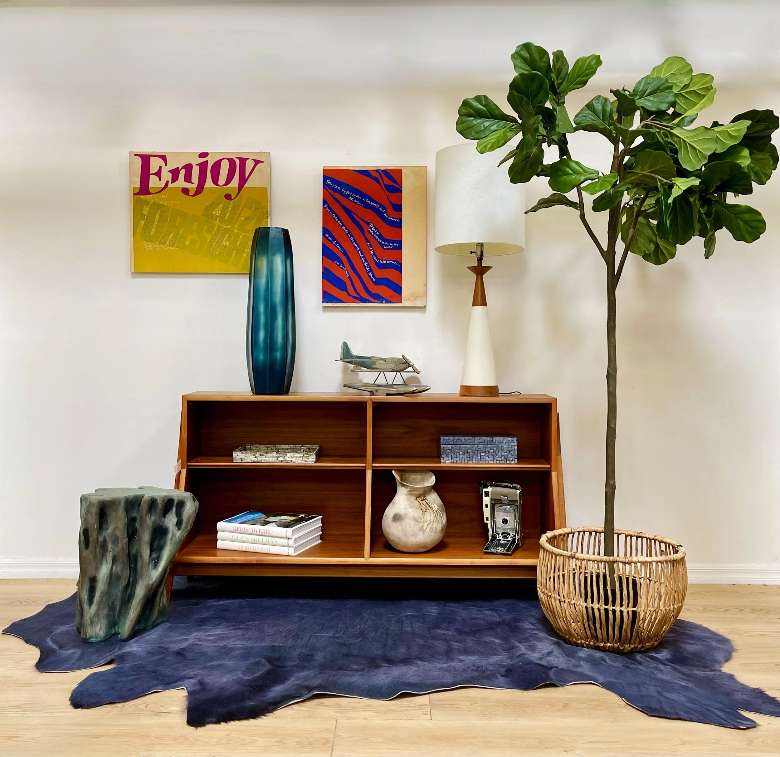 An eclectic collection of vintage and artistic items arranged on a wooden floor, including a carved totem pole, a painted canvas of a woman, a mannequin with a hat, a upholstered bench with a leather bag, an antique trunk, and a patterned rug.
