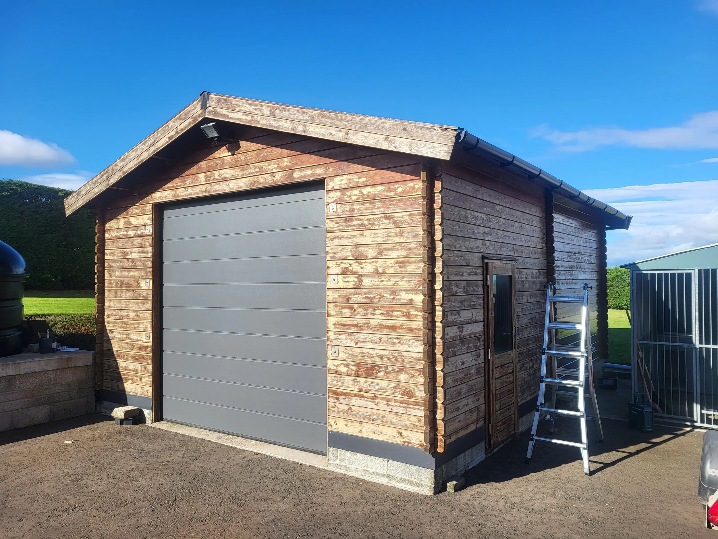 A small wooden building with a gray garage door, a sloped roof, and a ladder leaning against one side, outdoors on a sunny day with a clear blue sky.