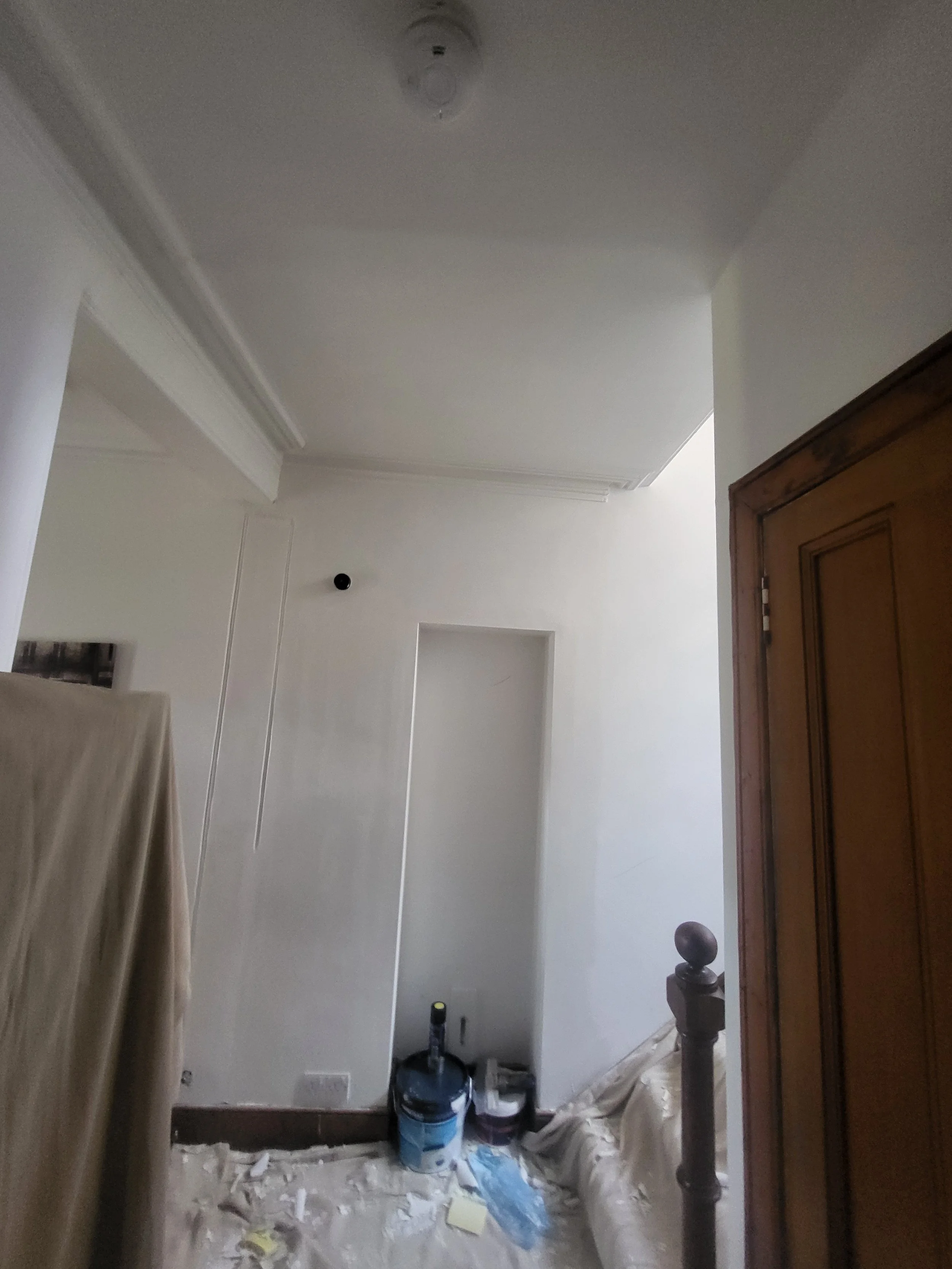 A partially painted room with painting supplies on the floor, a plastic drop cloth, paint cans, and a ladder in the corner.