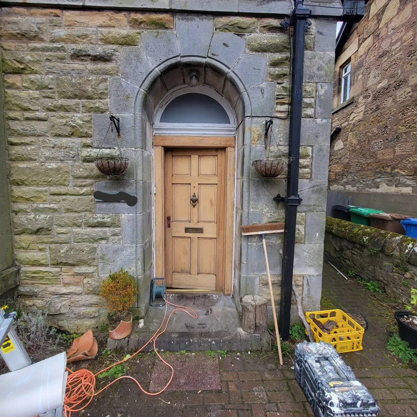 A small, rustic front door set in a stone wall, surrounded by gardening tools, a broom, a yellow crate, and various potted plants with bricks and outdoor furniture nearby.
