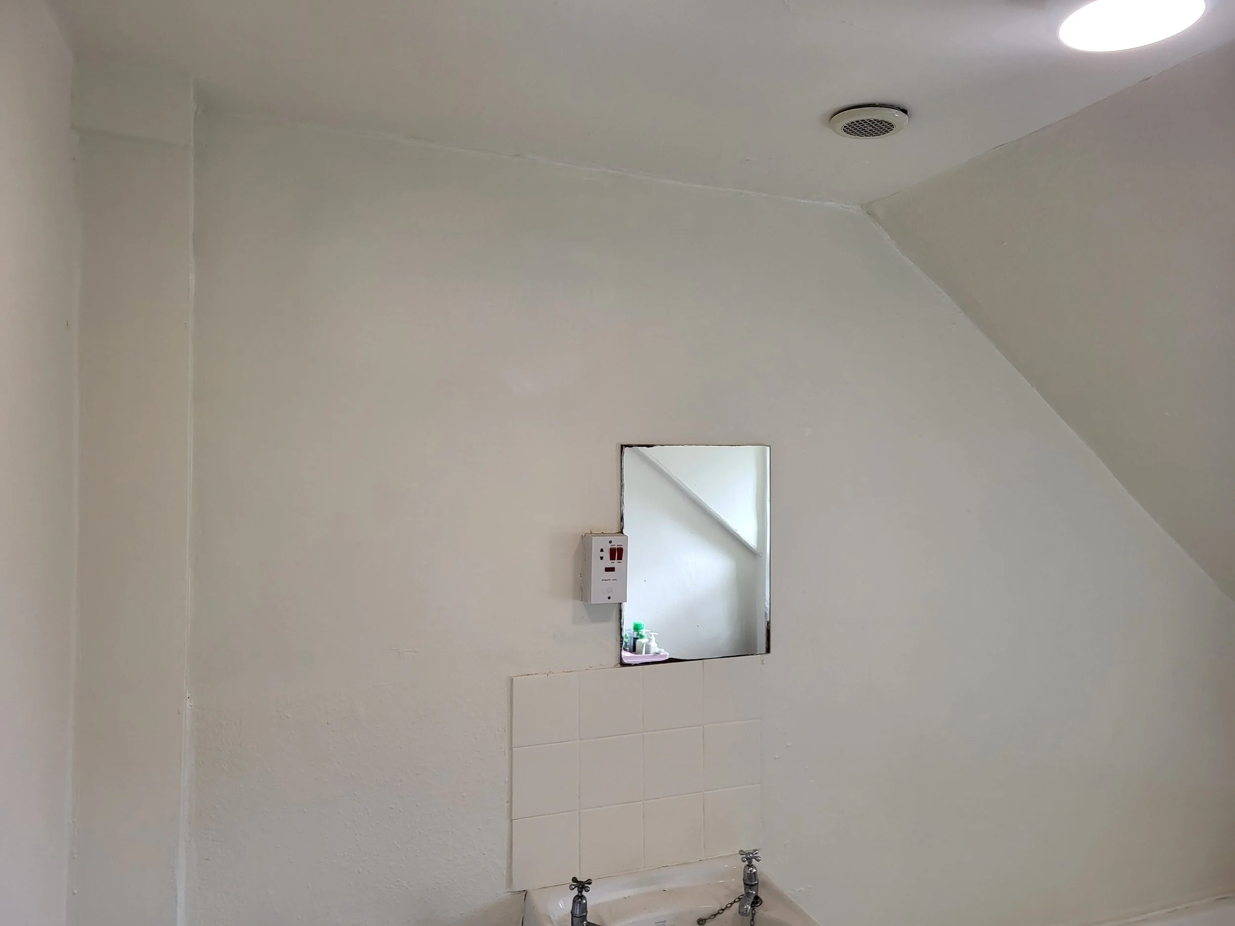 Empty room with white walls, ceiling, and tiles, a small square mirror on the wall, a window with ledge, and a white electrical outlet.