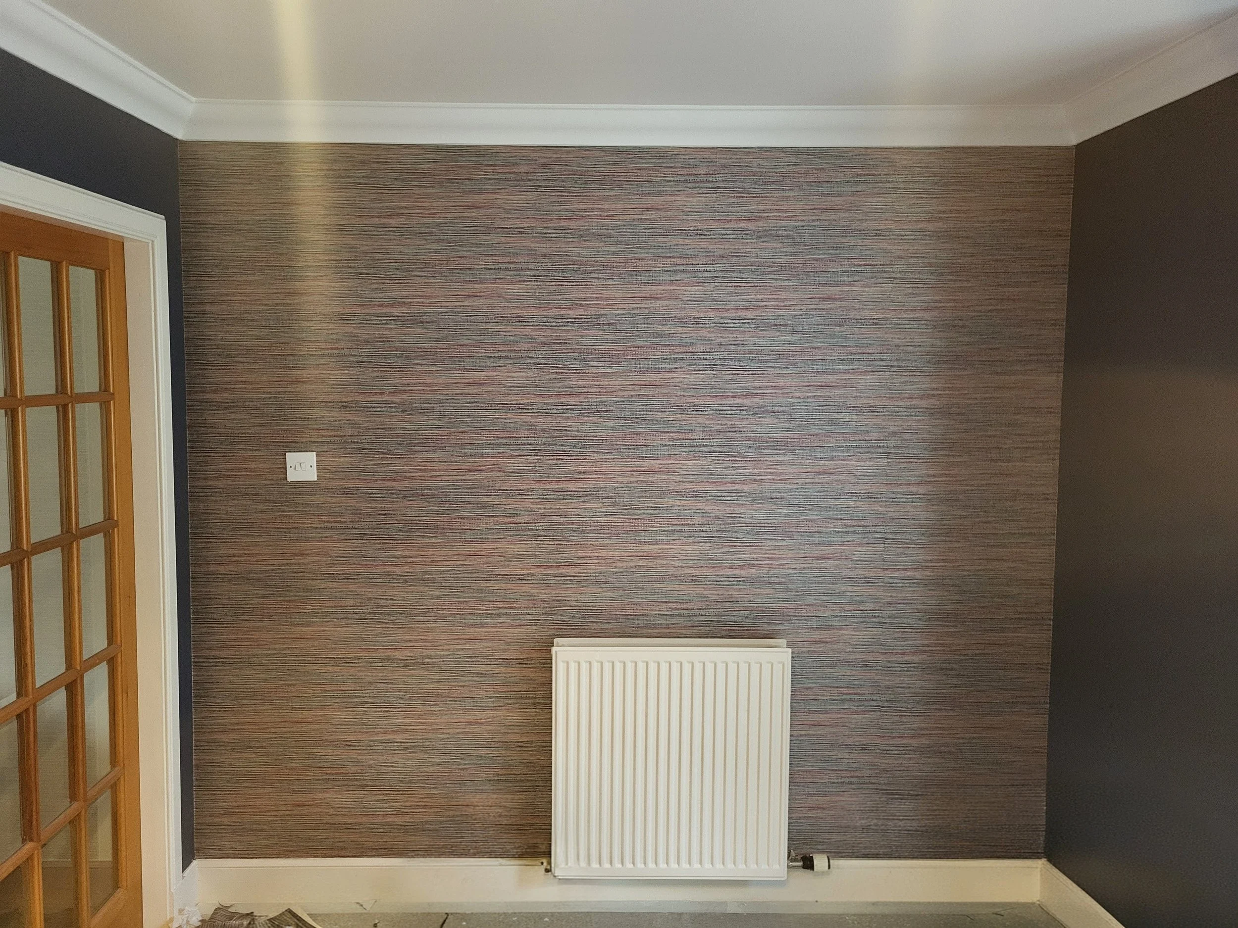 Interior view of a room with a wallpapered wall, a white radiator, a wooden door with glass panels, and a gray wall on the right.