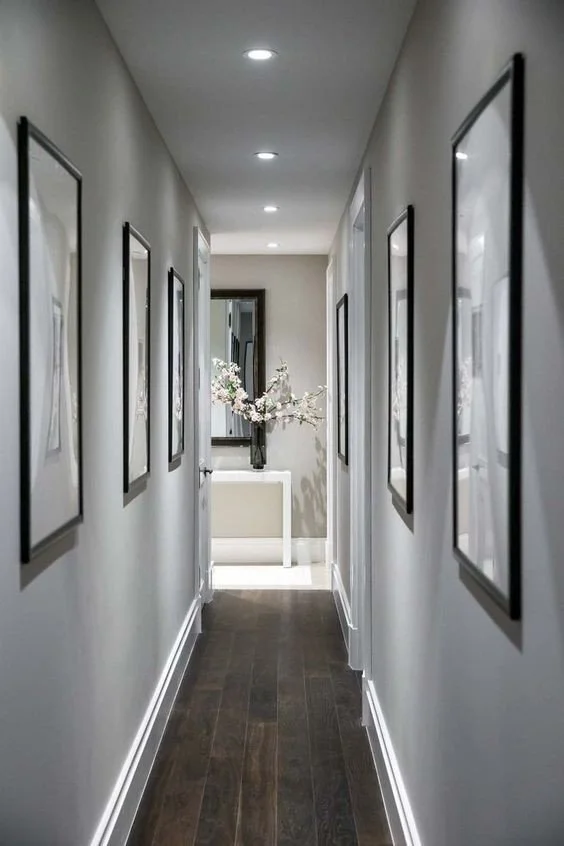 Narrow, well-lit hallway with framed artwork, dark wooden floor, and a console table with flower vase at the end.