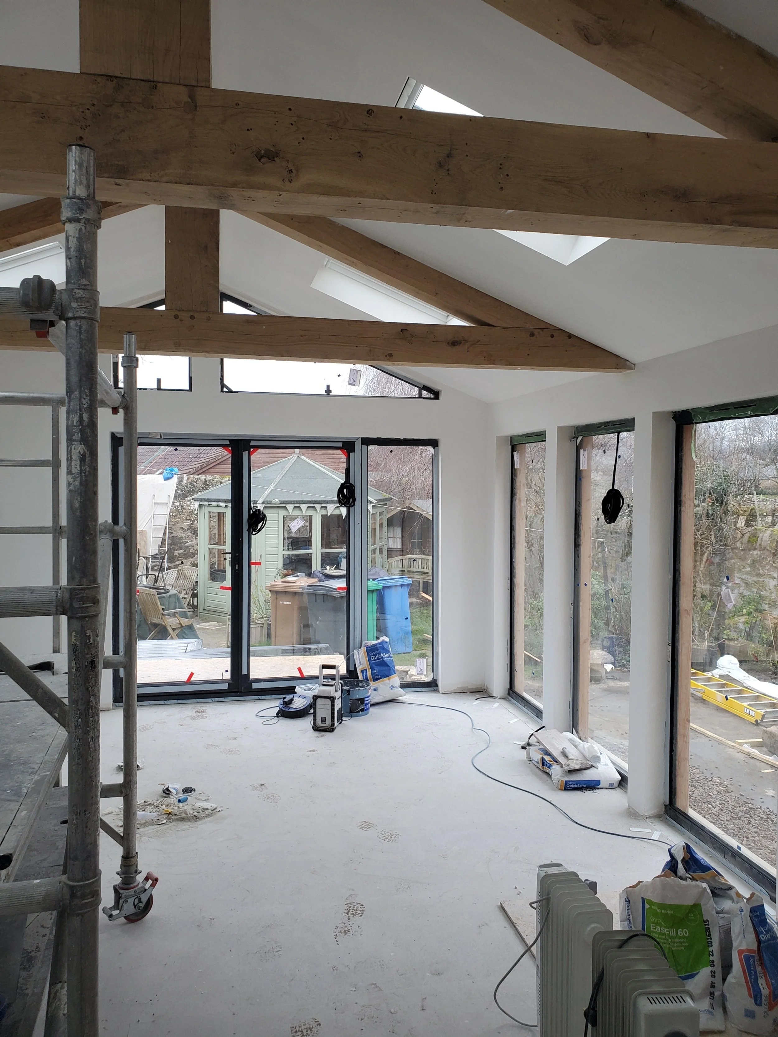 Interior of a room under renovation with exposed wooden beams, large windows, and construction tools scattered around.