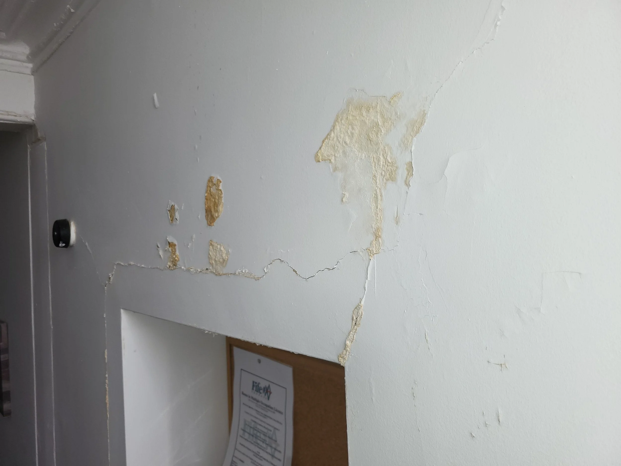 Close-up of a damaged wall with peeling paint and cracks, a sign in a frame is partially visible on the lower part of the image.