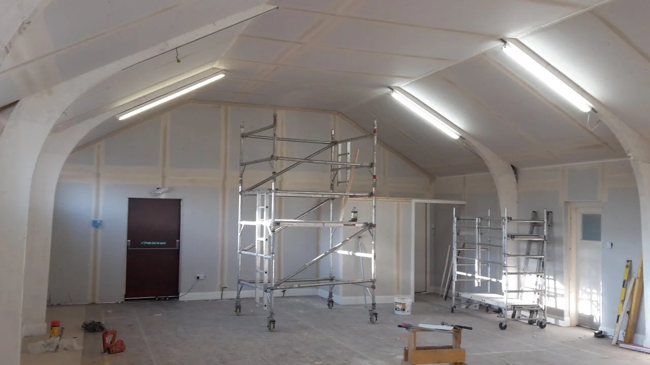 Interior under construction with scaffolding and tools, unfinished walls, and ceiling lights.