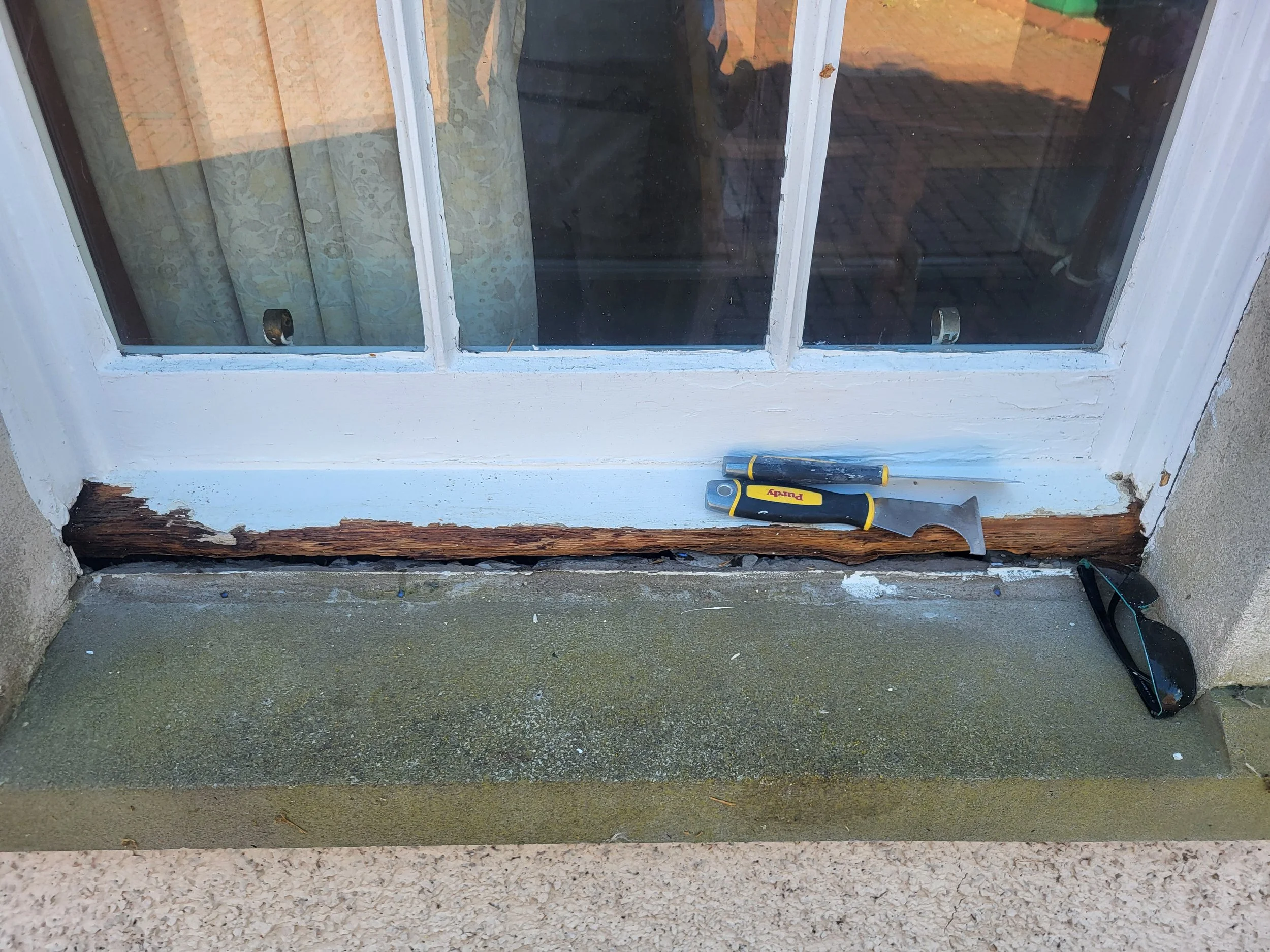 Window with white frame being repaired, with tools like a utility knife placed on the ledge; the window has a damaged wooden sill with peeling paint and exposed wood.