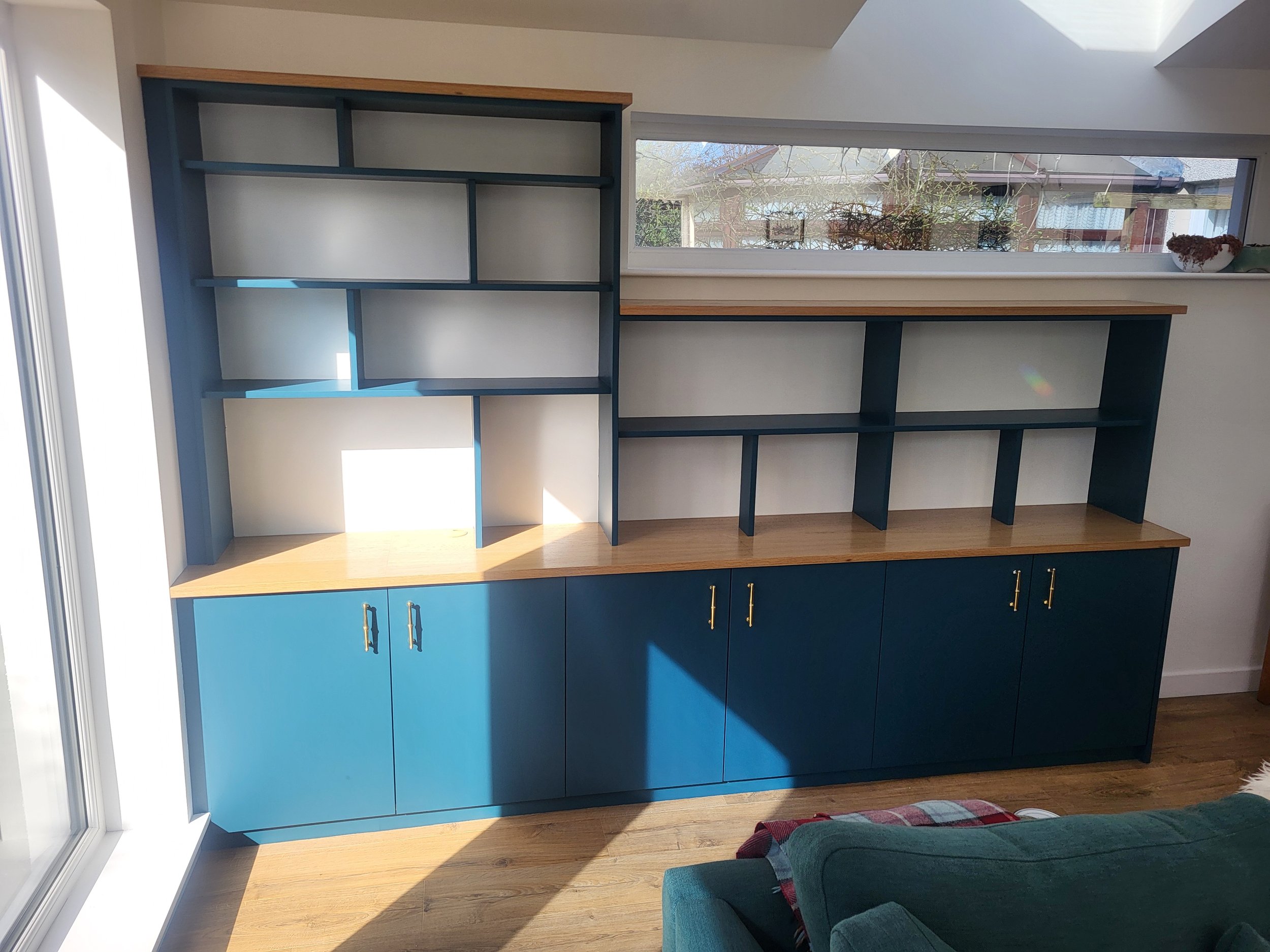 A modern interior room features blue cabinets with gold handles, a wooden top shelf, and a wall-mounted wooden shelf unit painted in blue. There is a large window letting in sunlight and a view of outside houses and trees. A colorful armchair is part