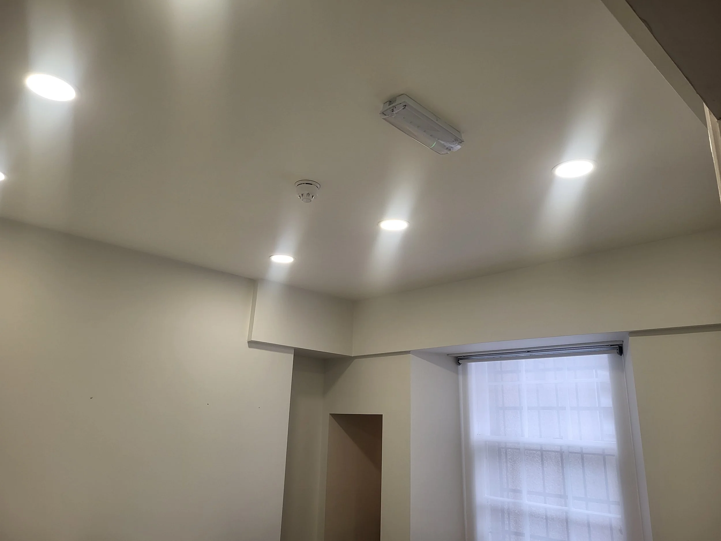 The ceiling of a room with four recessed lights, a smoke detector, and a mounted electrical fixture, with a window visible in the corner.