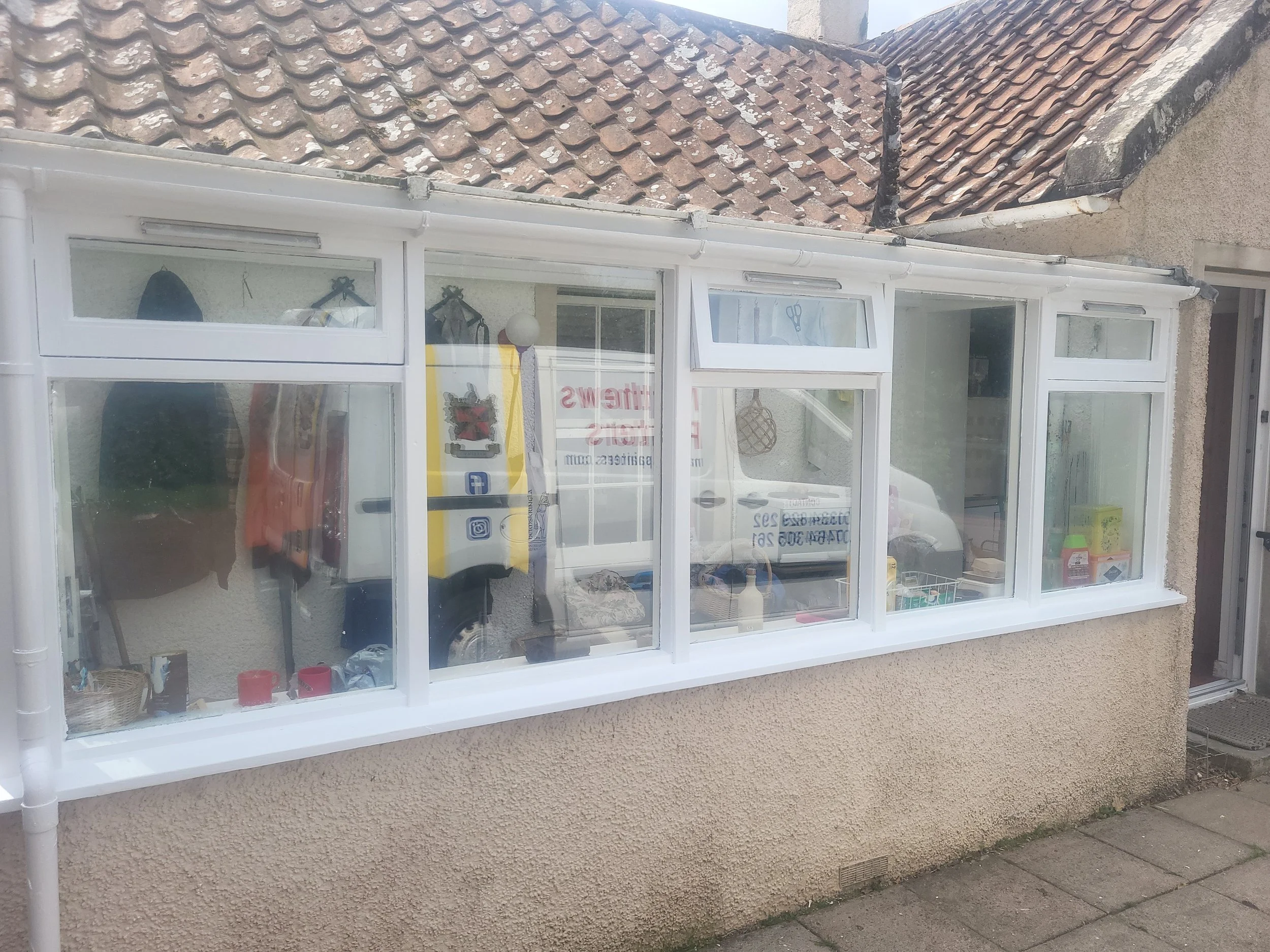 A house window with white framing and glass panes, displaying some items inside including a yellow sign, clothes hanging, and various small objects. The house has a textured beige exterior wall, and the roof above is made of reddish-brown tiles with some white patches. An outside drain is also visible at the bottom of the wall.