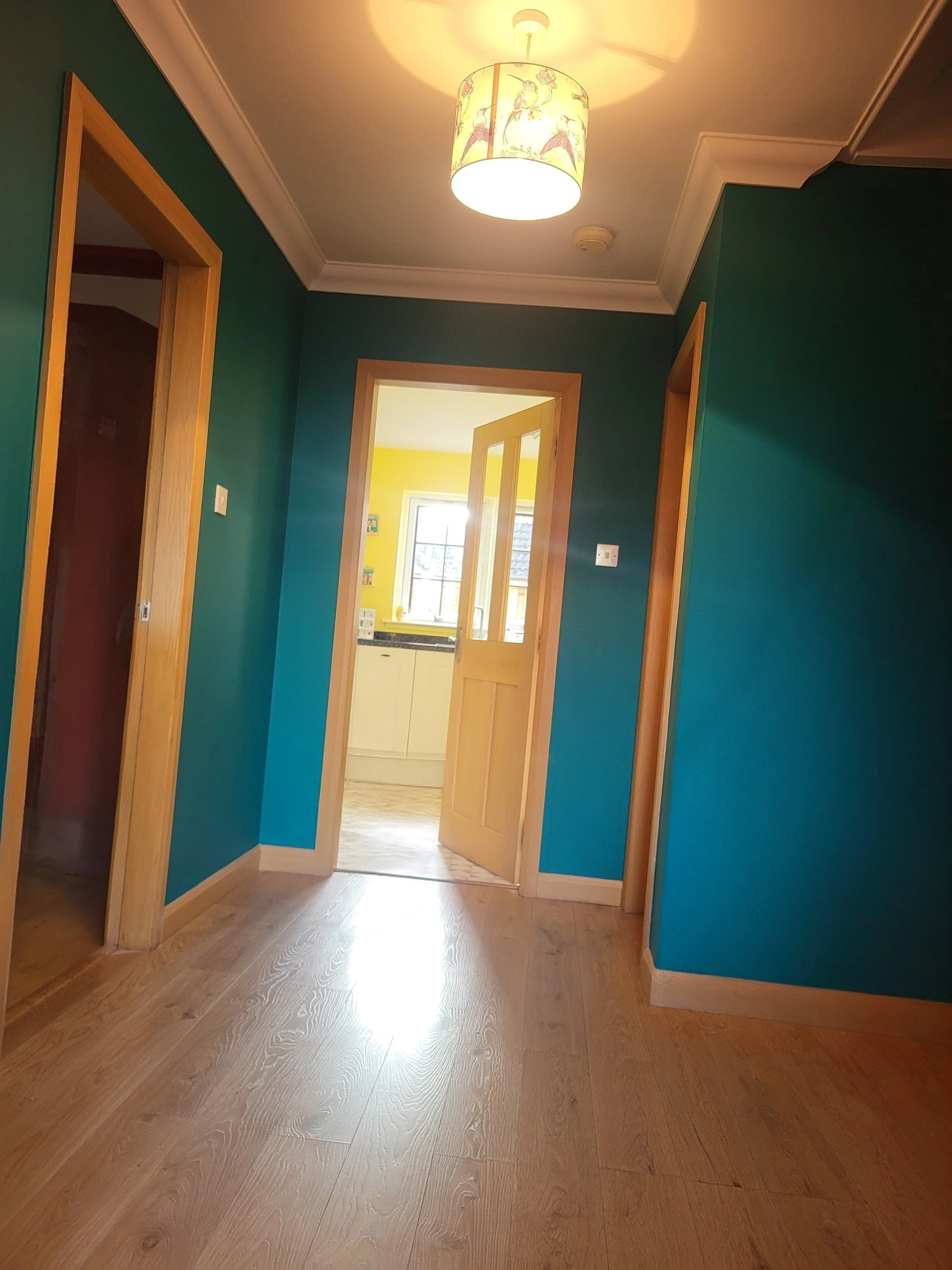 Interior view of a hallway with teal walls, wooden door frames, and a ceiling lamp, leading to a yellow-colored kitchen in the background.