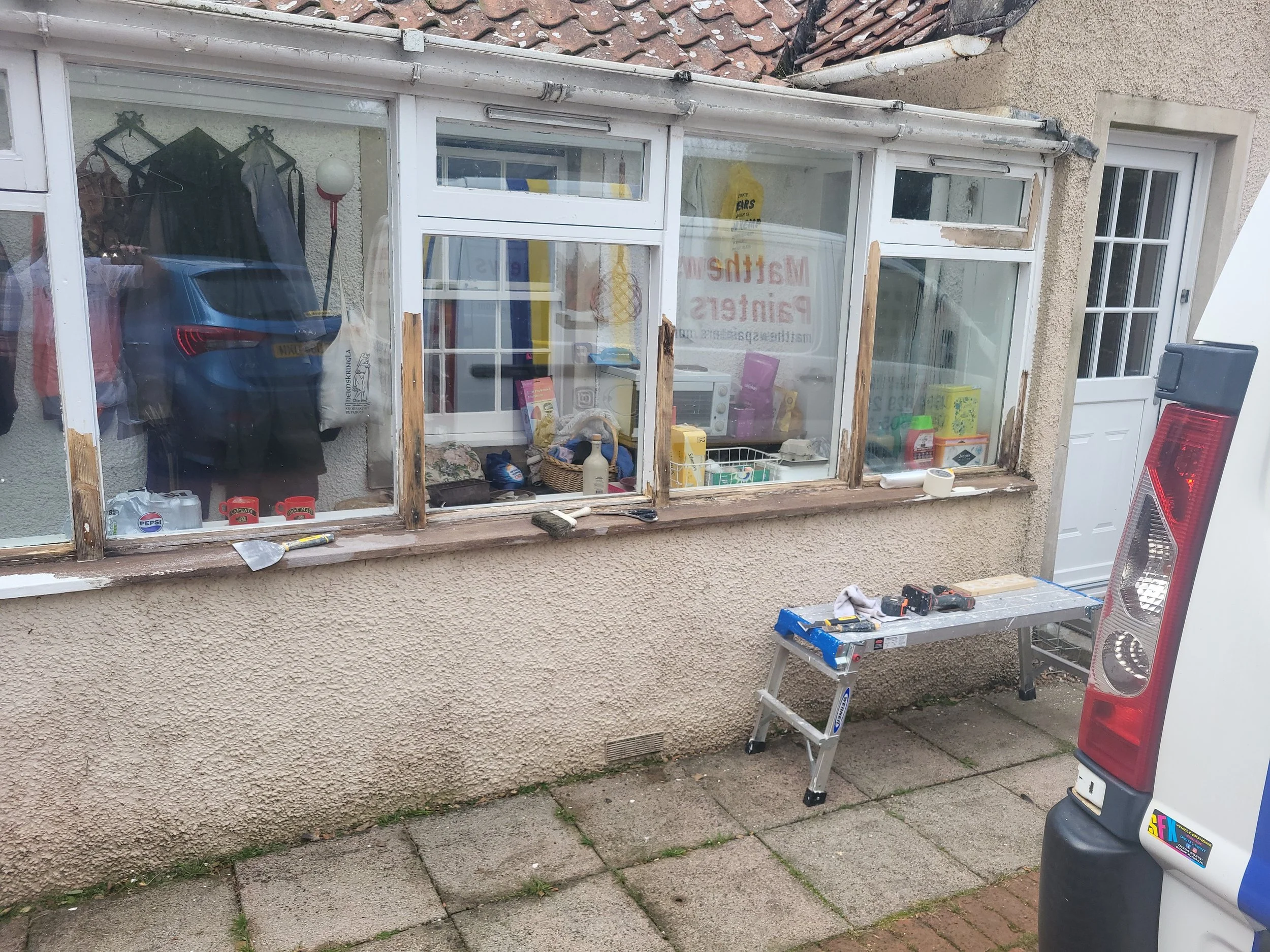 A house with a large front window that is being renovated, with tools and supplies outside, including a step ladder, and a mirror of old tiles and patches of new wood around the window frame.