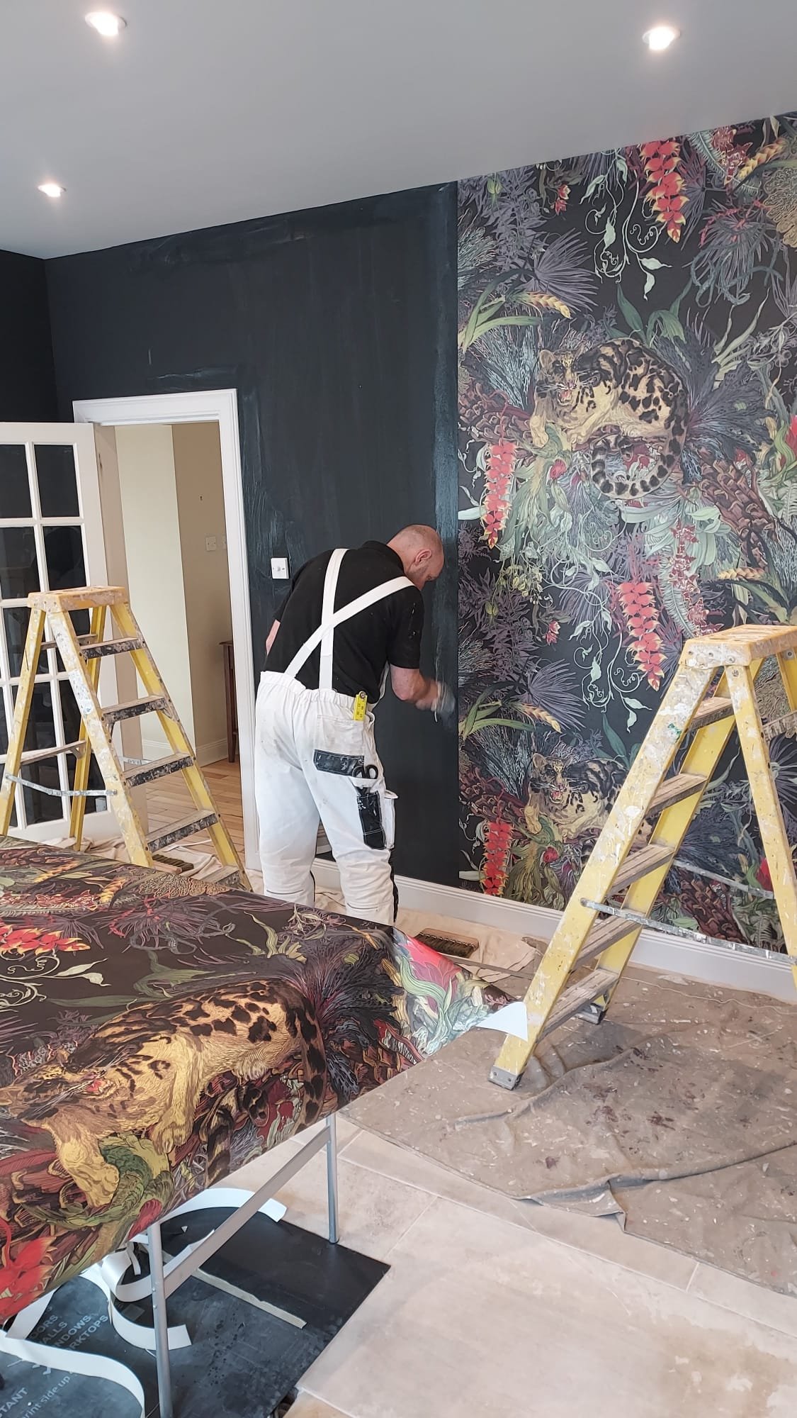 A person painting or applying wallpaper on a dark wall in a room, with a large floral and animal mural on an adjacent wall, and two ladders present.