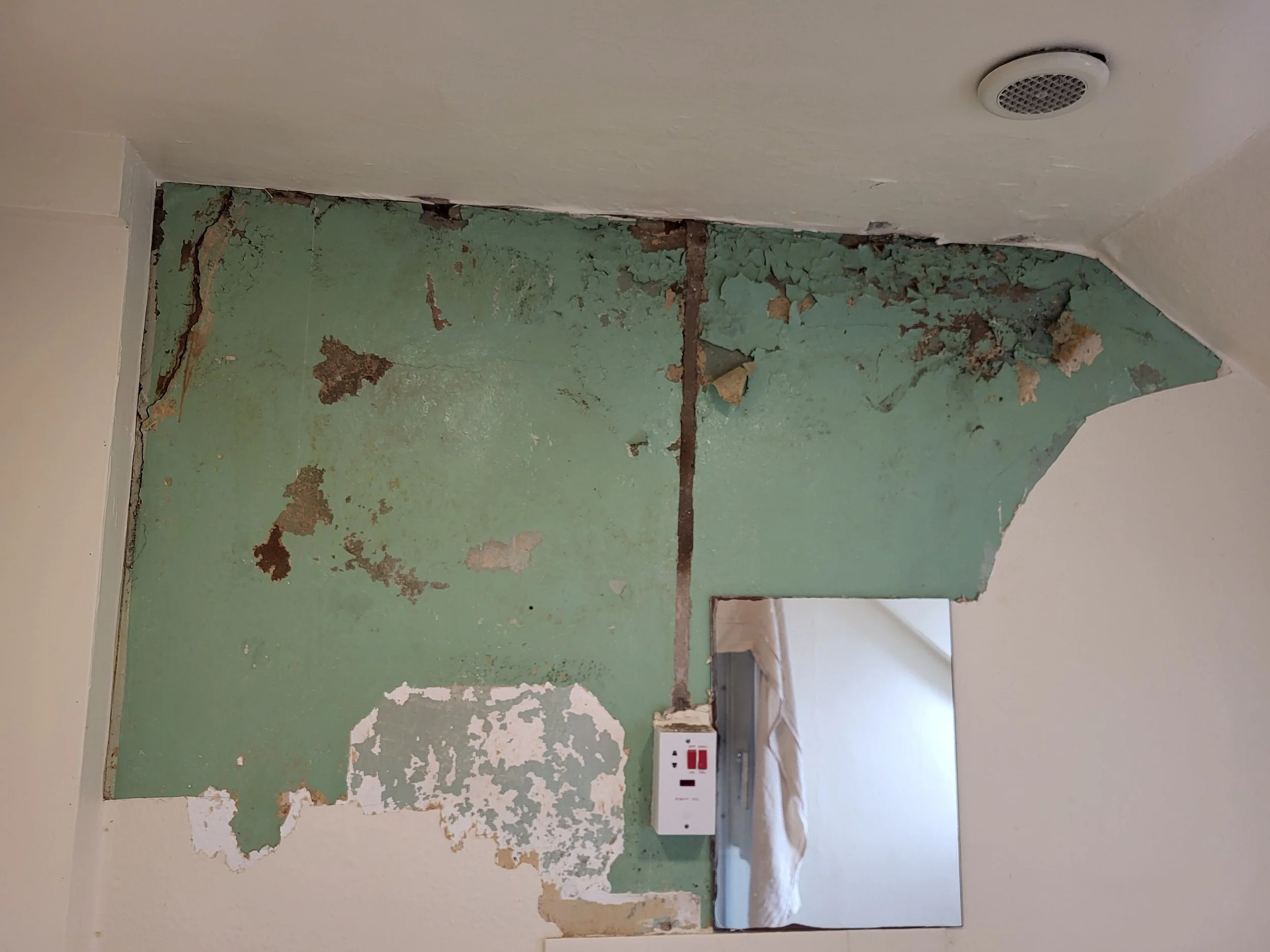 A partially demolished green wall with peeling paint and exposed drywall, with a white outlet and a covered electrical cable on the floor.