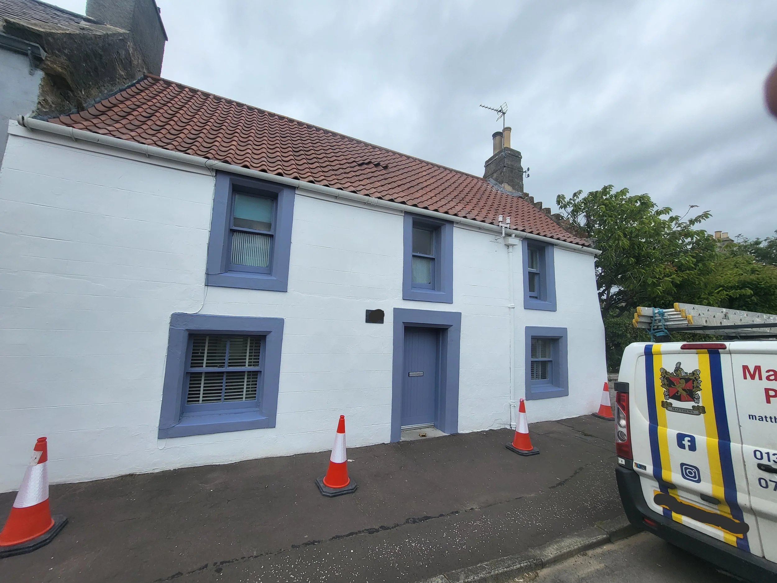 A house with white walls, purple window frames, and a red-tiled roof, with construction cones and a service van parked in front.