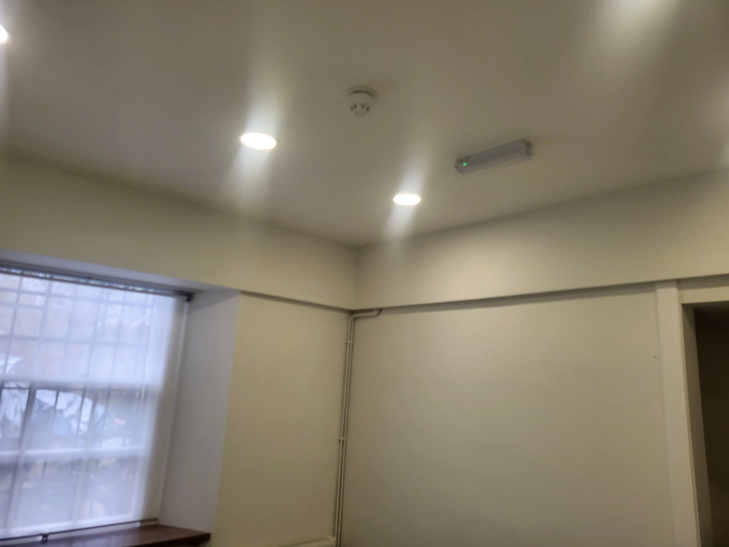 The image shows a room with a ceiling, recessed lights, a smoke detector, and a mounted electronic device with a green indicator light. There is a window with blinds on the left side.