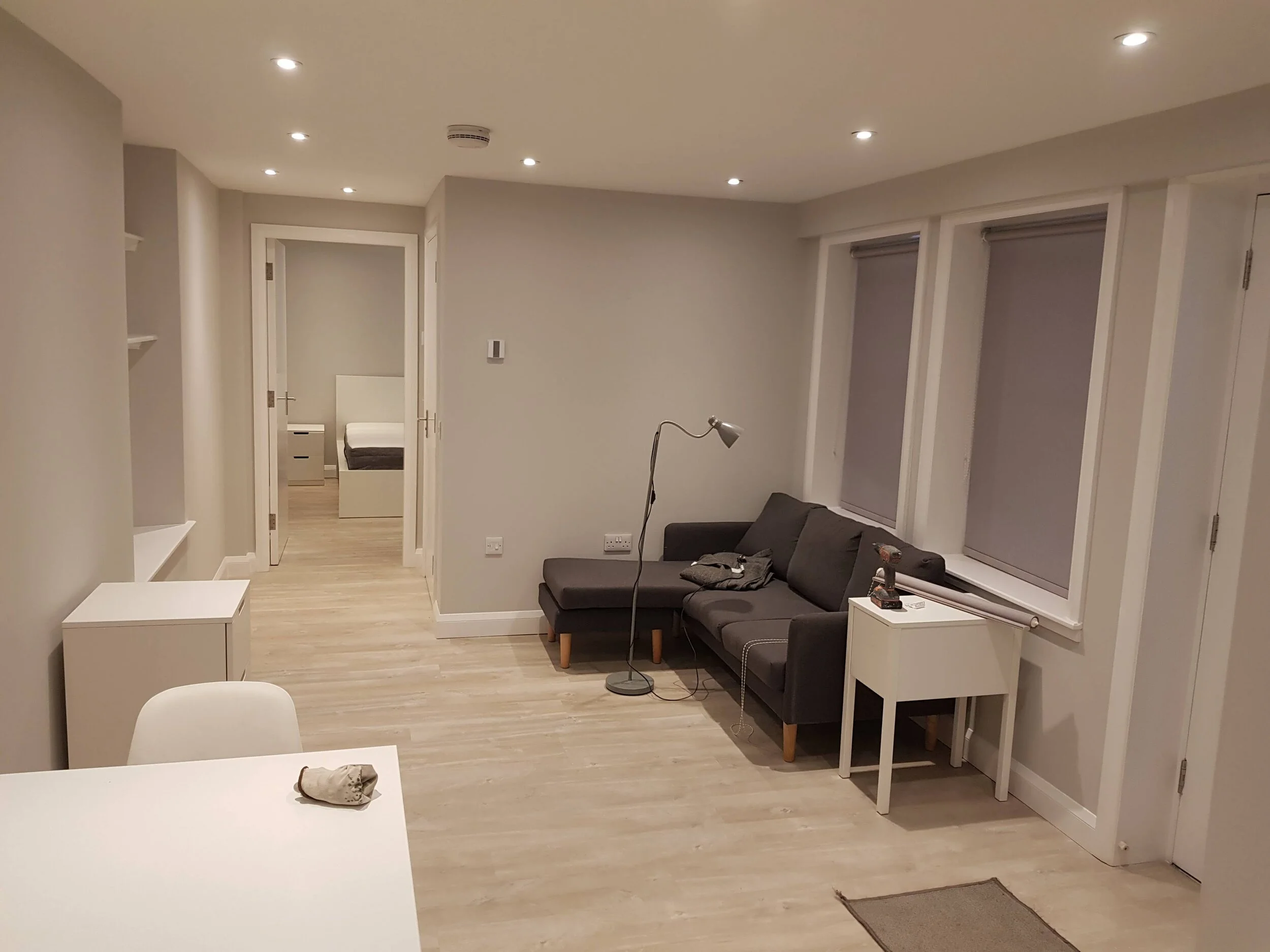 Modern living room with a gray sofa, white side tables, floor lamp, and a view into a bedroom. Room is minimally decorated with wooden flooring and recessed lighting.