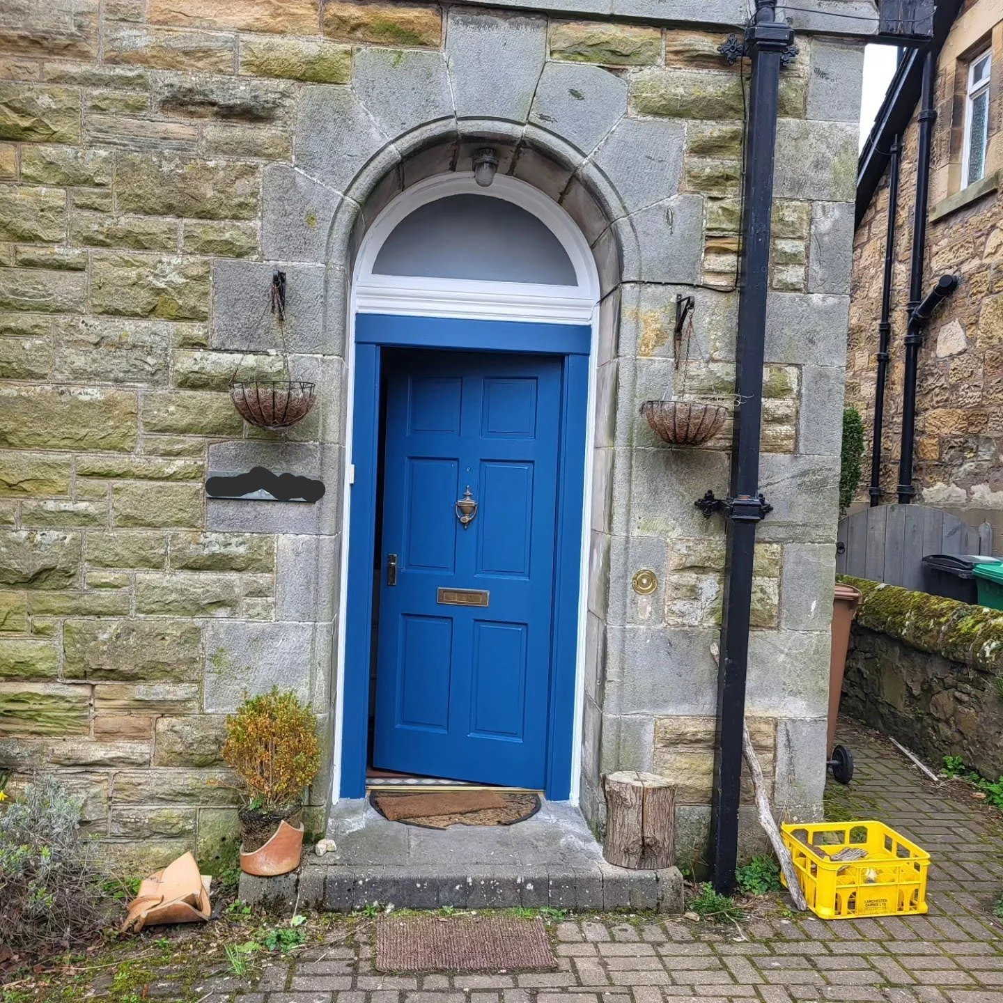Stone house entrance with a blue door, arched above, with a brass letterbox and door knocker. Two hanging baskets are on either side of the door. Small shrubs and a log are near the door. To the right, black drain pipes run down the wall, with a yellow crate on the ground.
