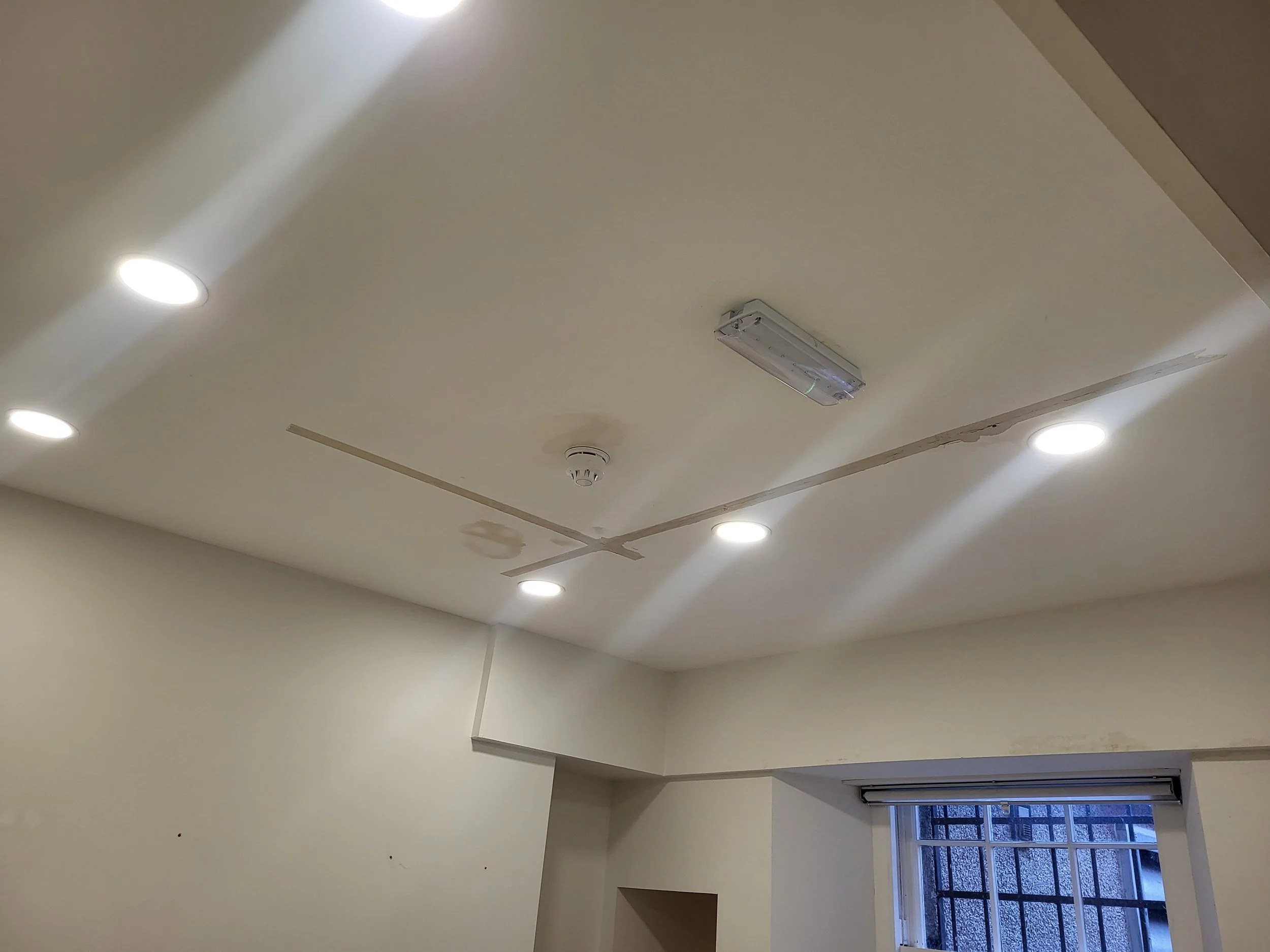 The image shows a ceiling with four recessed lights, a smoke detector, and a rectangular ceiling vent. Part of a window with a metal grate is visible at the bottom right corner.