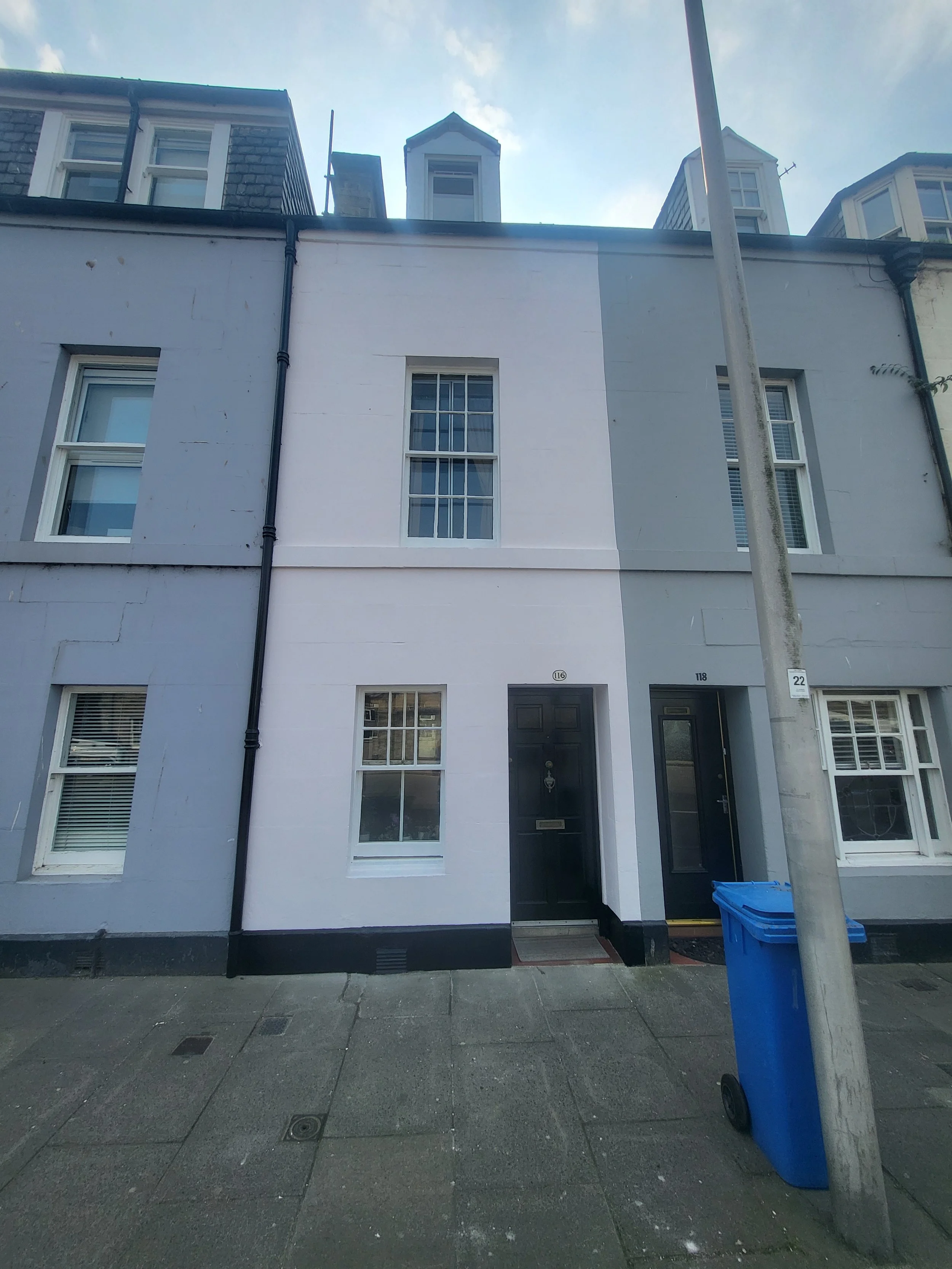 Front view of two adjacent middle-row townhouse units with dark doors, white and grey facades, a blue trash bin, and a streetlamp in front.
