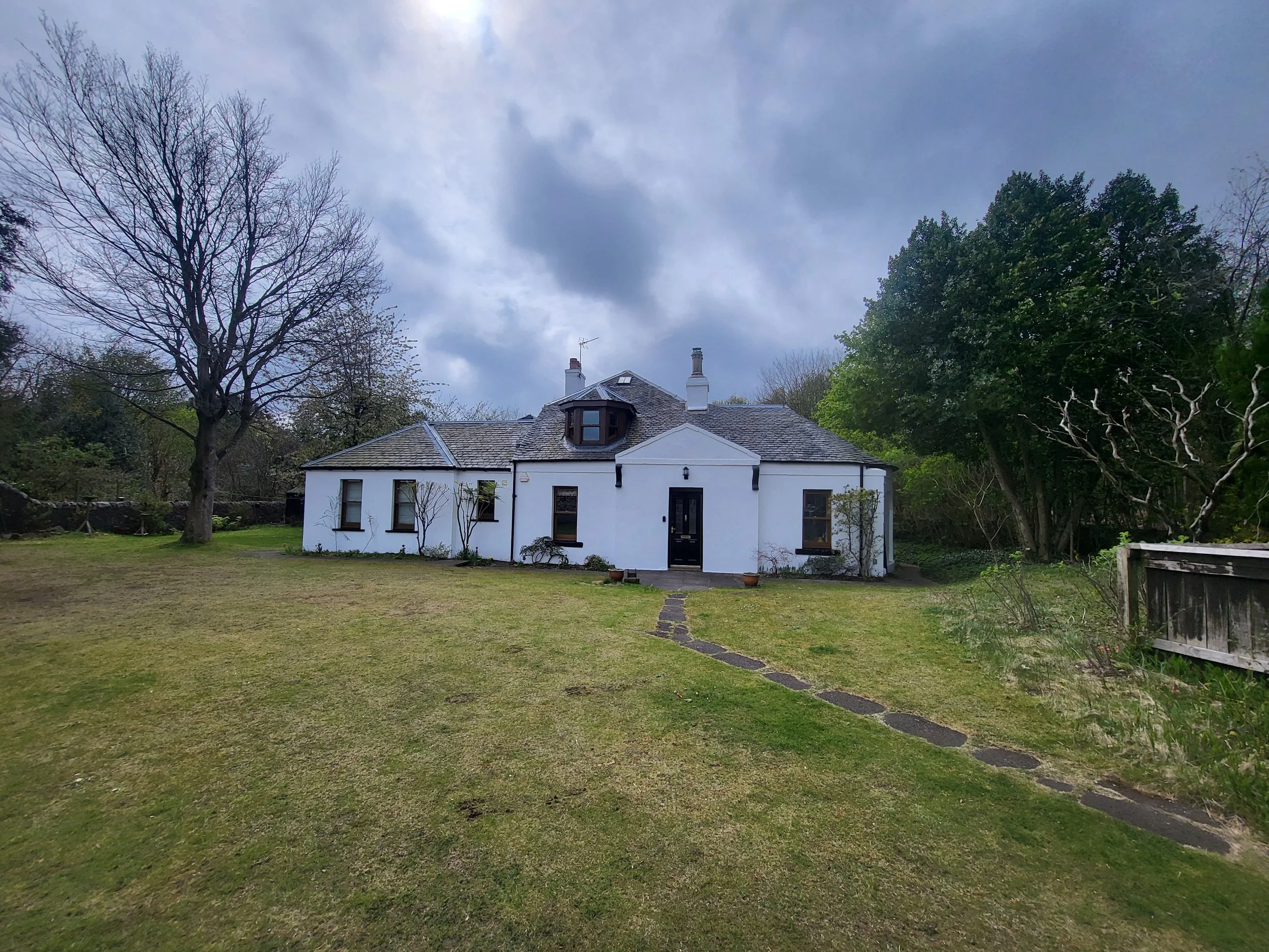 White house with black door, situated in a grassy yard with a stone pathway leading to the entrance, surrounded by trees under a cloudy sky.