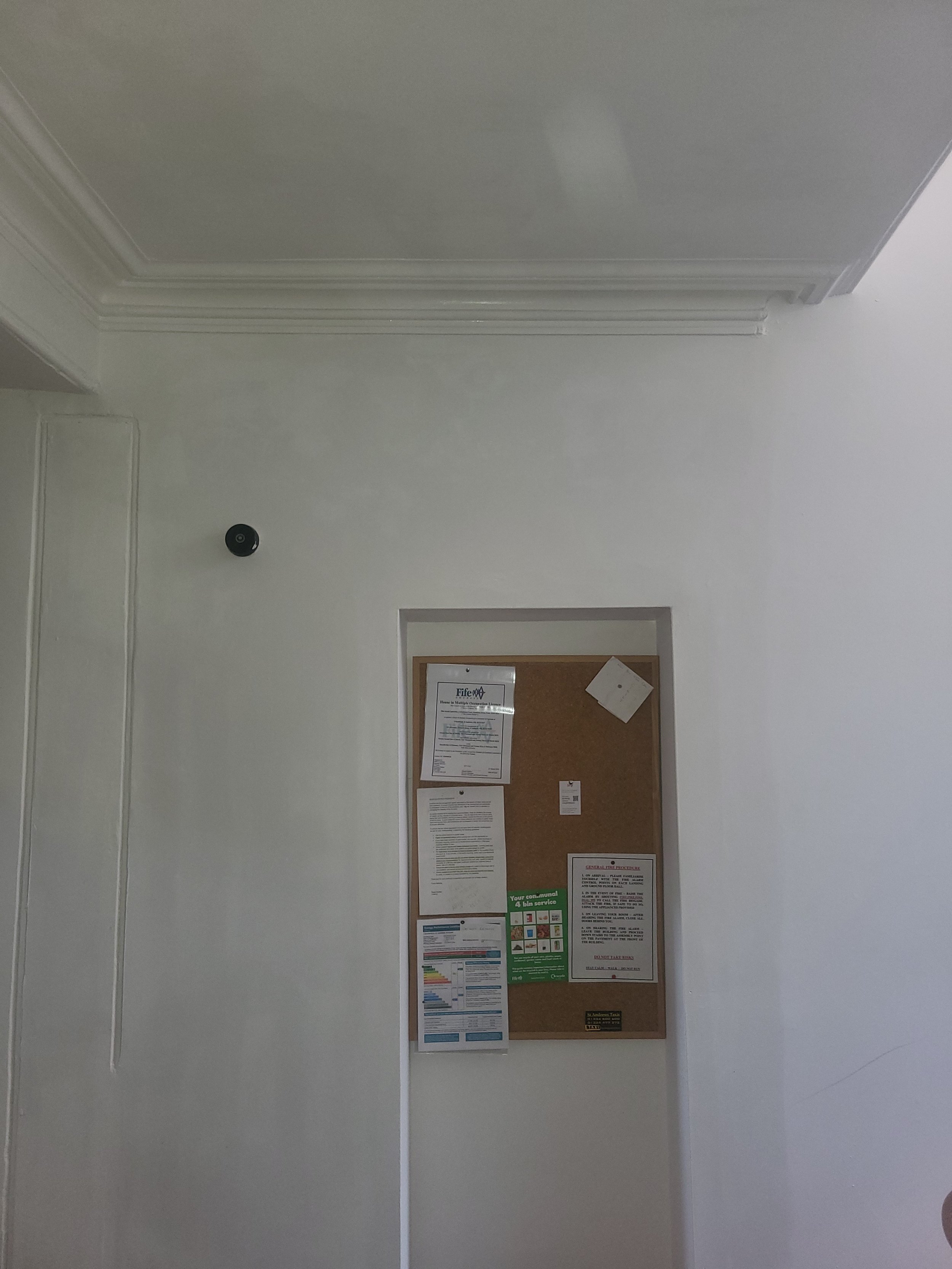 A bulletin board with printed notices and a small white paper posted on a white wall, with a doorway and a black alarm or security camera on the wall nearby.