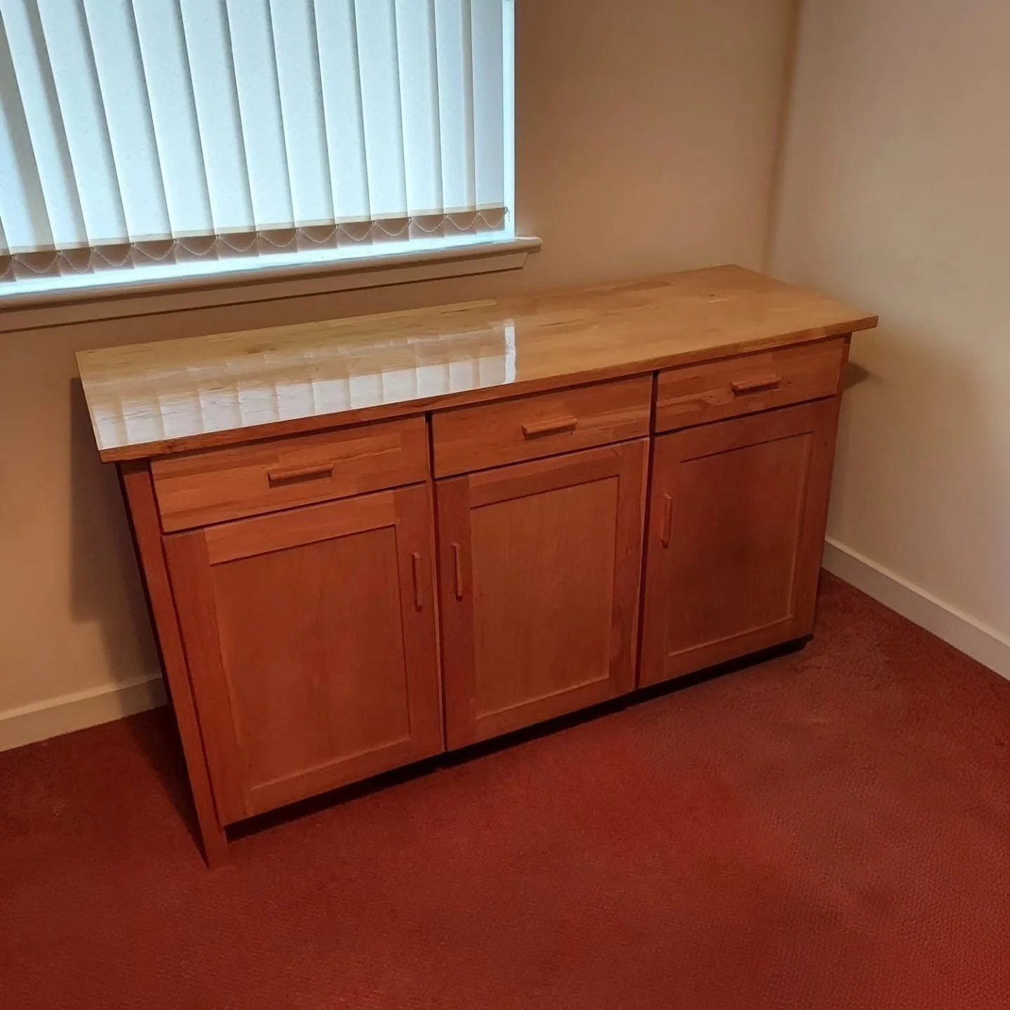 A wooden sideboard with three doors and two drawers, placed against a beige wall under a window with closed vertical blinds, on a reddish-brown carpeted floor.