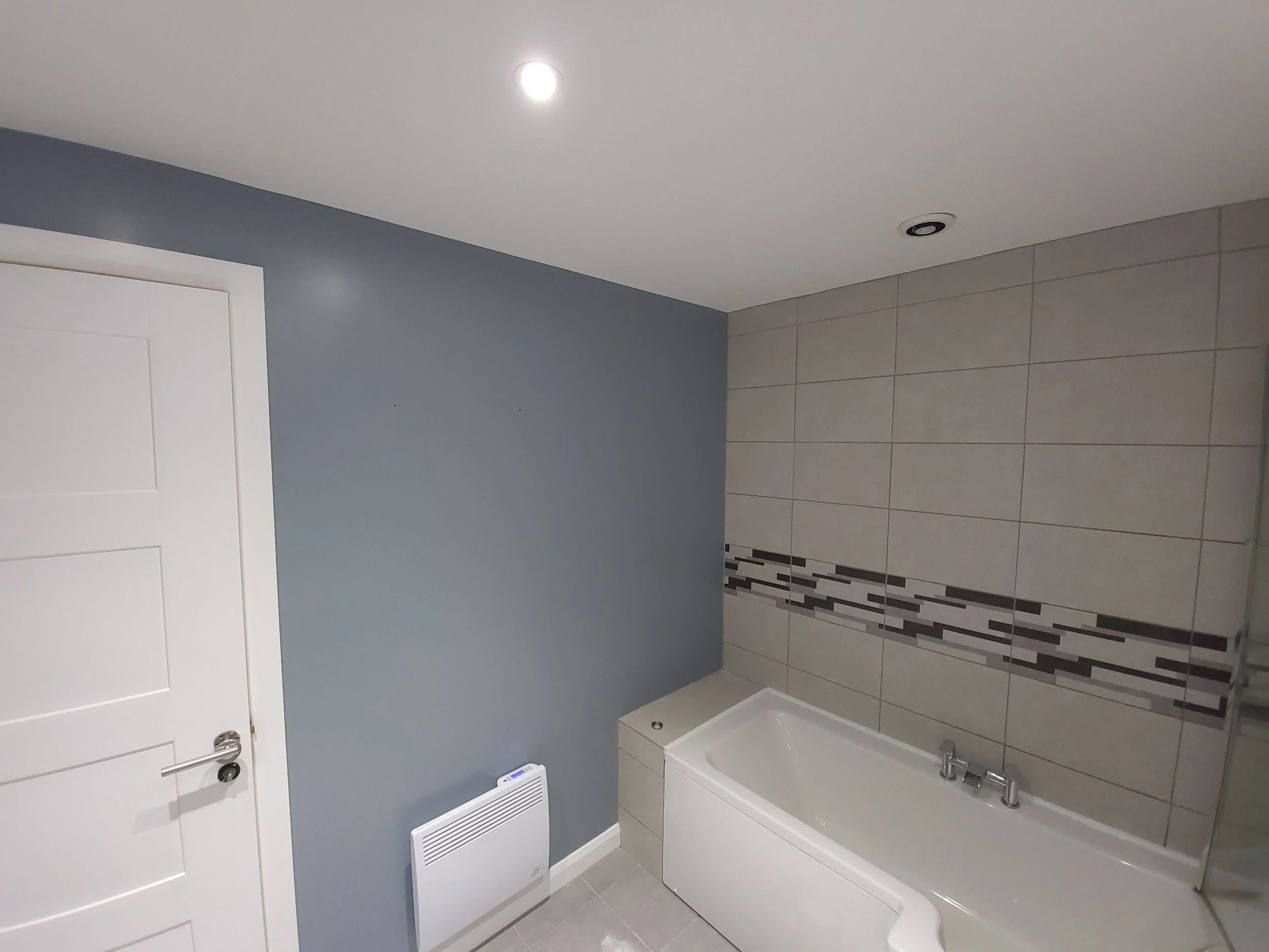 Modern bathroom interior with white bathtub, gray tile wall, white door, blue wall, and ceiling light.