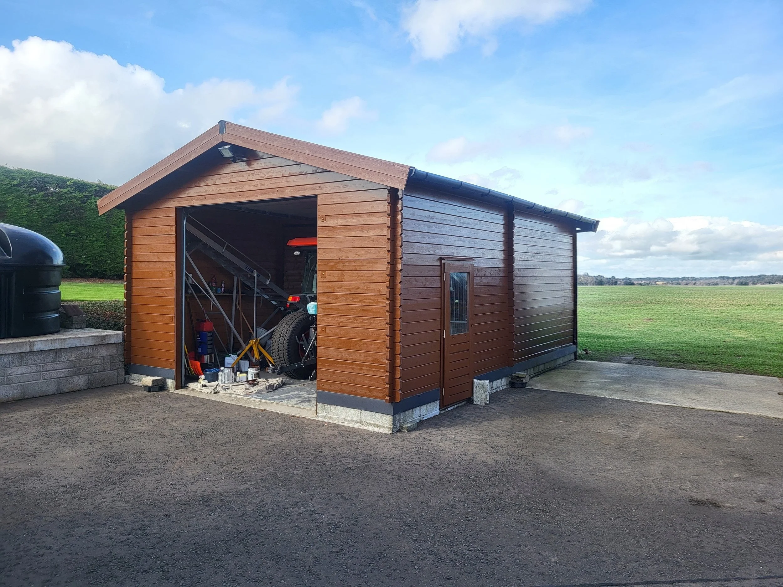 A wooden shed with an open door, filled with various tools and equipment, situated on a concrete and gravel surface with a grassy field and blue sky with clouds in the background.