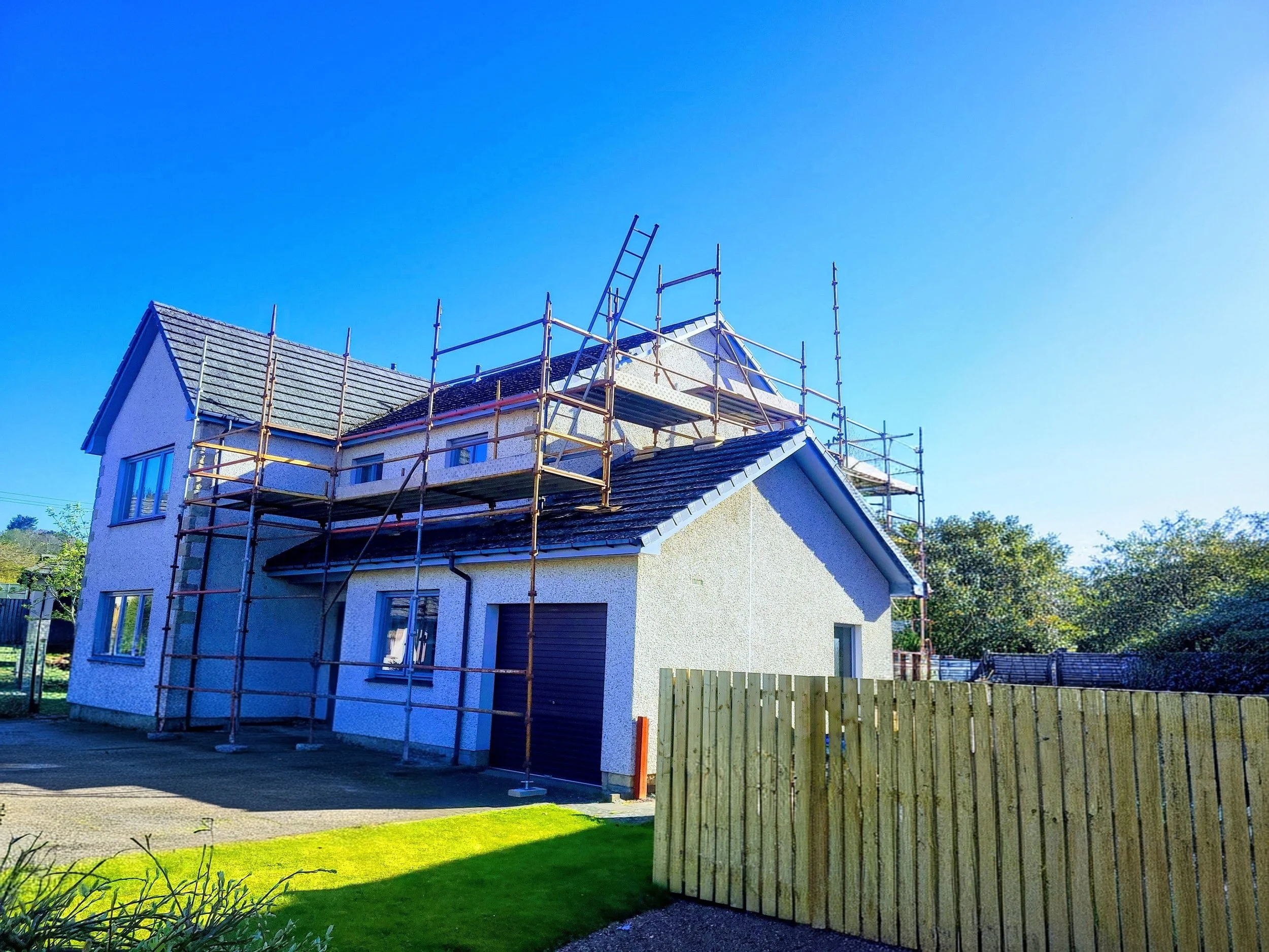 A house with scaffolding around the roof, appearing to be under construction or renovation, with clear blue sky in the background.