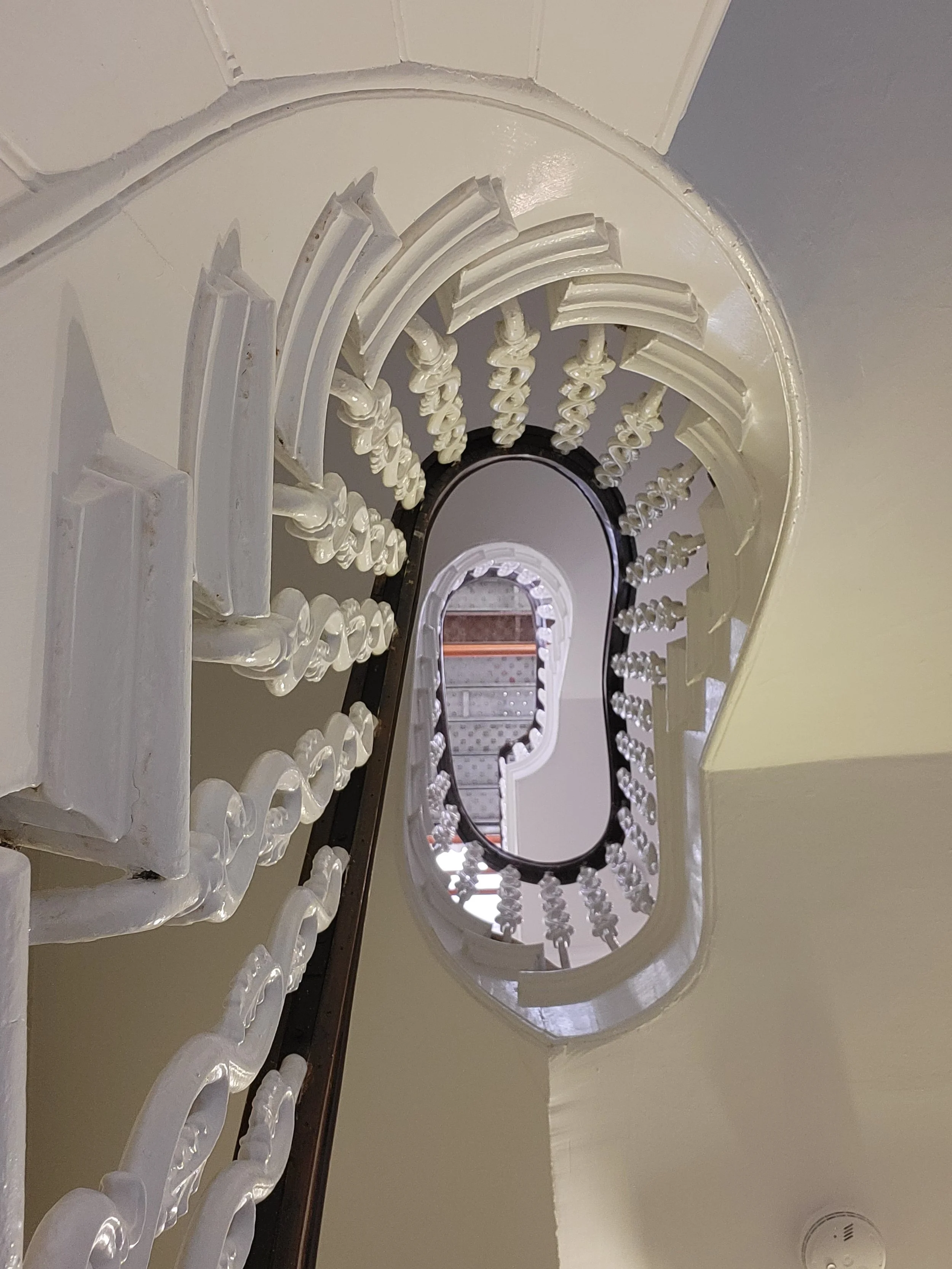 Looking up at a spiral staircase with ornate white balusters and a wooden handrail, viewed from the ground floor.