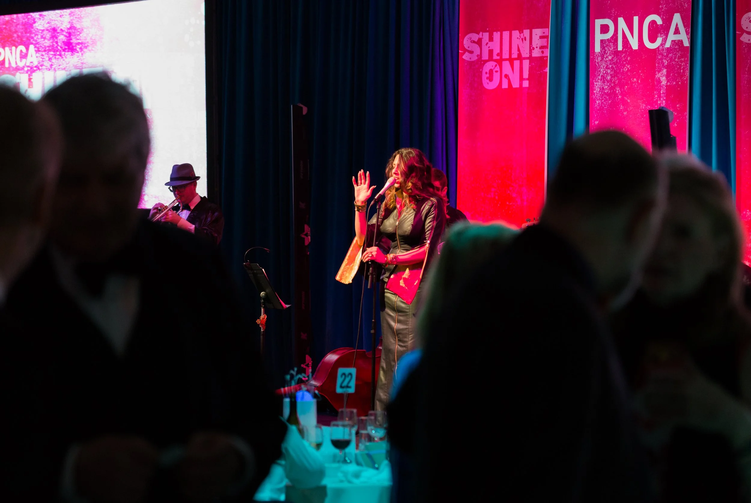 A singer on stage performing at an event with the words 'Shine On!' and 'PNCA' on pink background banners, audience members in formal attire, some wearing face masks, some drinking wine.