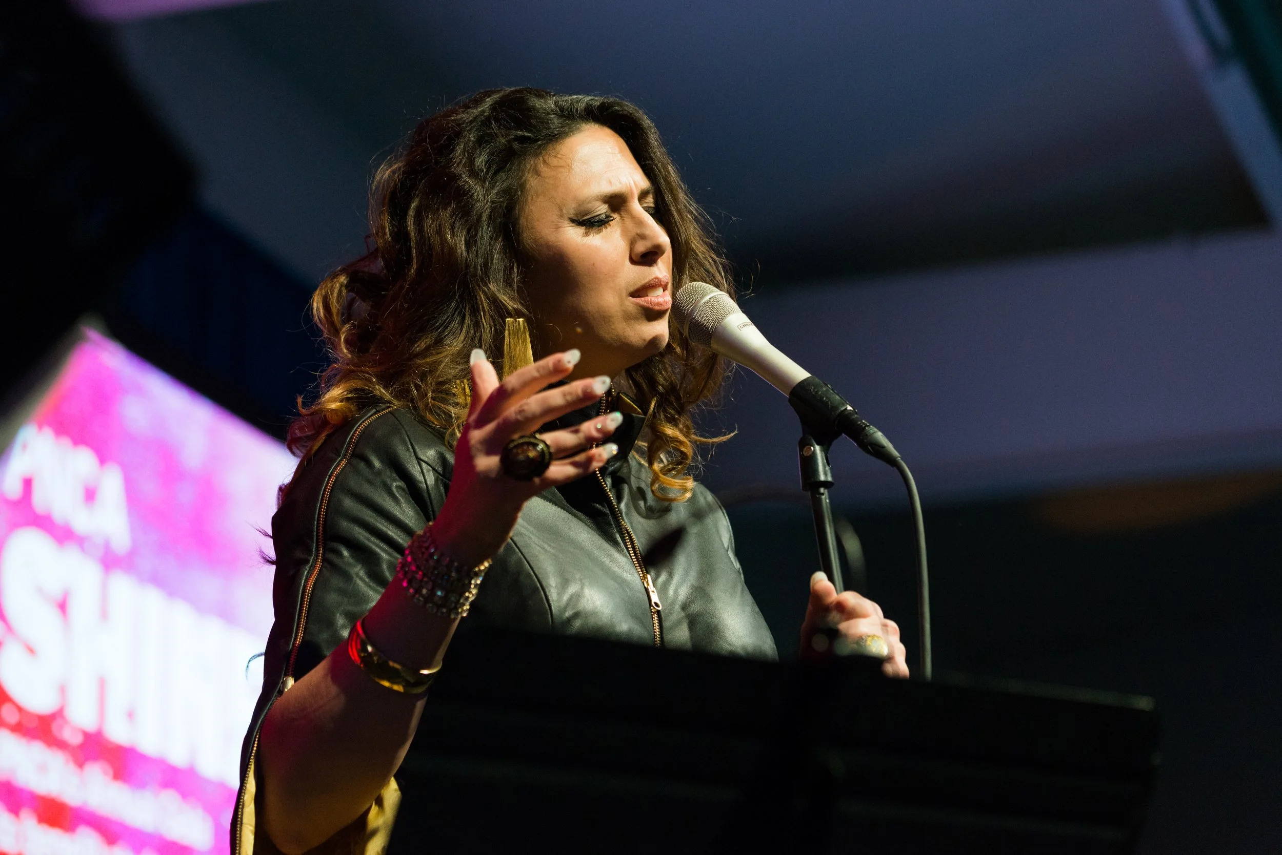 Tasha with curly hair singing into a microphone on a stage, wearing a leather jacket and jewelry.
