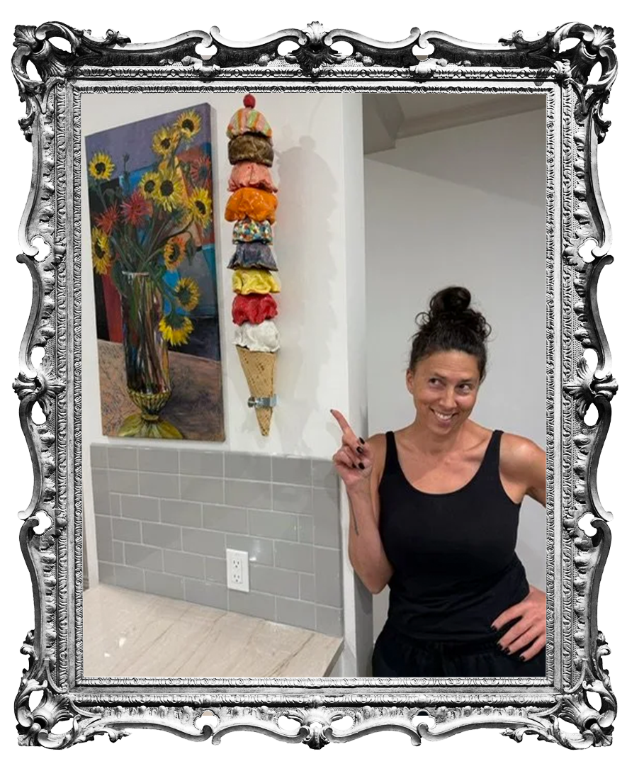 A woman in a black tank top with her hair in a bun standing in a room next to a wall with gray tiles, a painting of yellow and red flowers in a vase, and a vertical sculpture of stacked colorful clothing and an ice cream cone. The woman is smiling and pointing upward with her index finger.