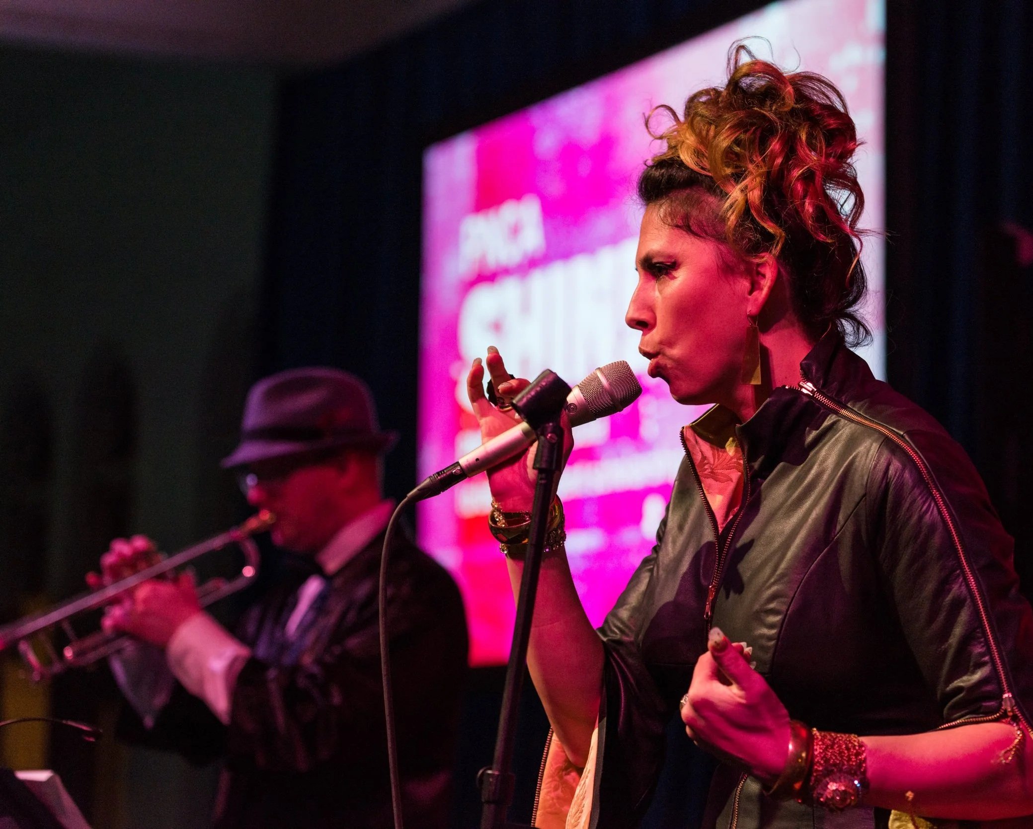 Tasha sings into a microphone with a man playing the trumpet in the background. Both are performing on stage with a purple and pink background.