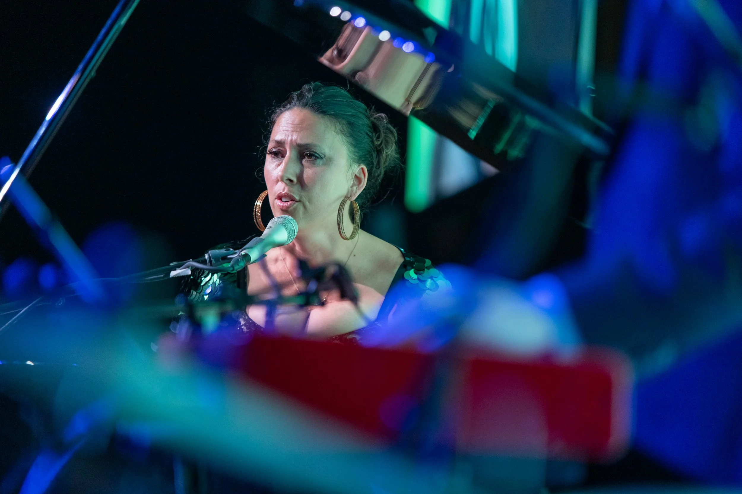 Tasha singing into a microphone at a piano during a performance, illuminated by blue and green stage lights.
