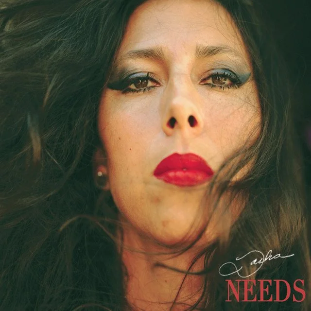 Close-up image of a woman with dark hair, bold makeup including blue eyeshadow, thick eyeliner, and bright red lipstick, with the text "asha NEEDS" in the bottom right corner.