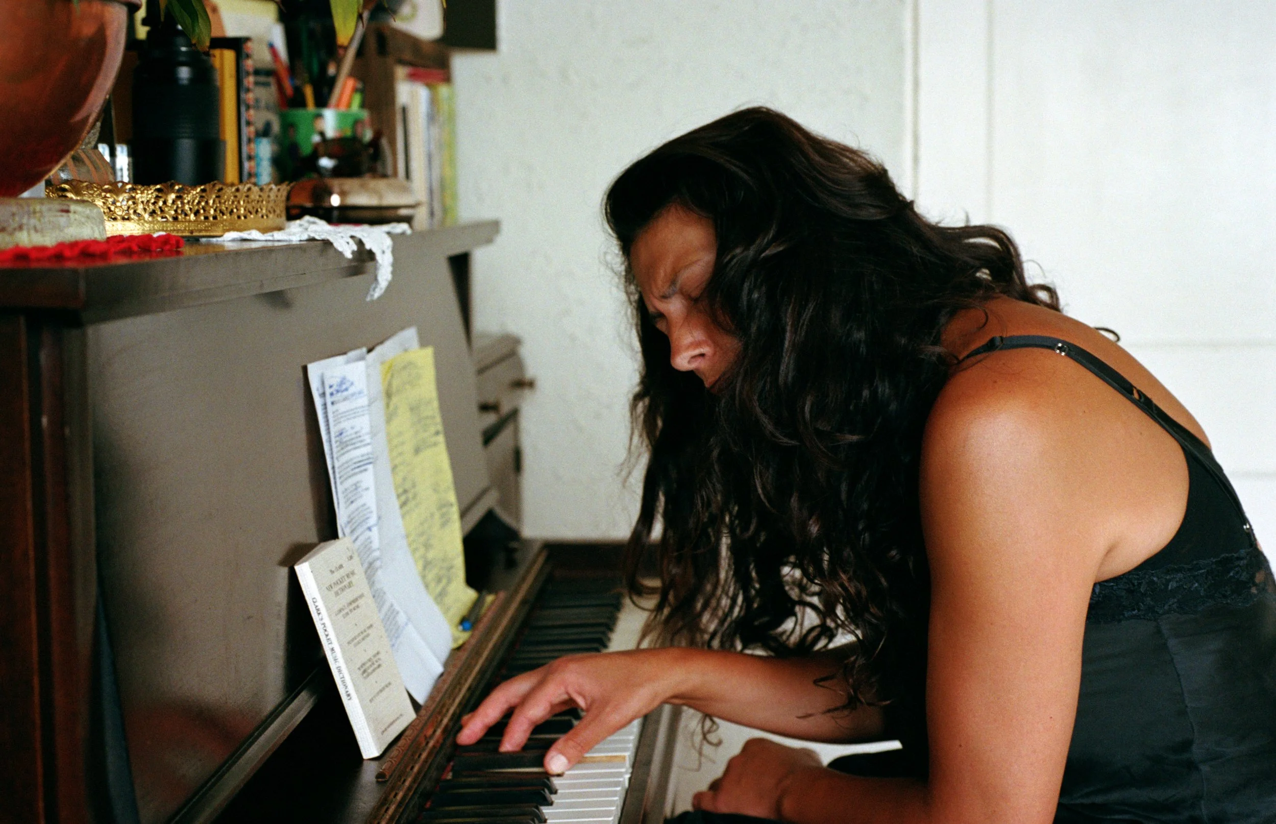 Tasha with long curly dark hair is playing the piano, with her head bent forward, wearing a black sleeveless top.