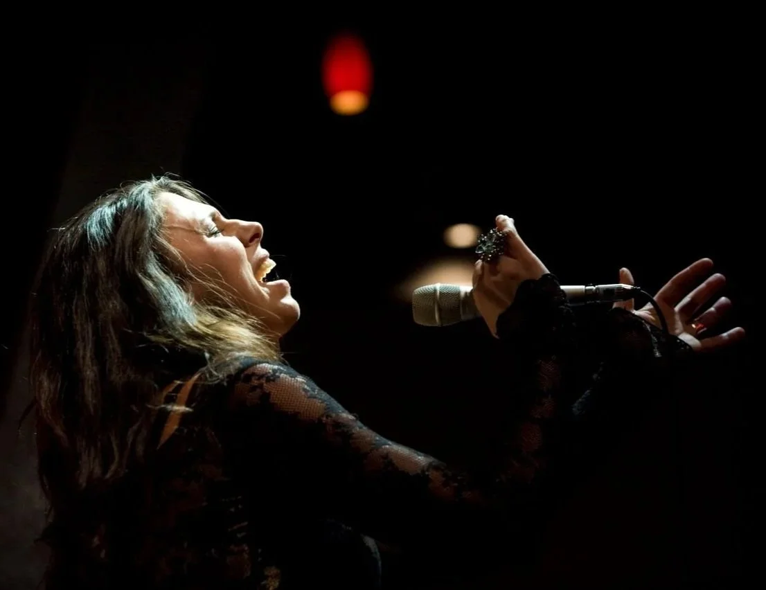 Tasha singing passionately into a microphone on a dark stage, with a red hanging light above.