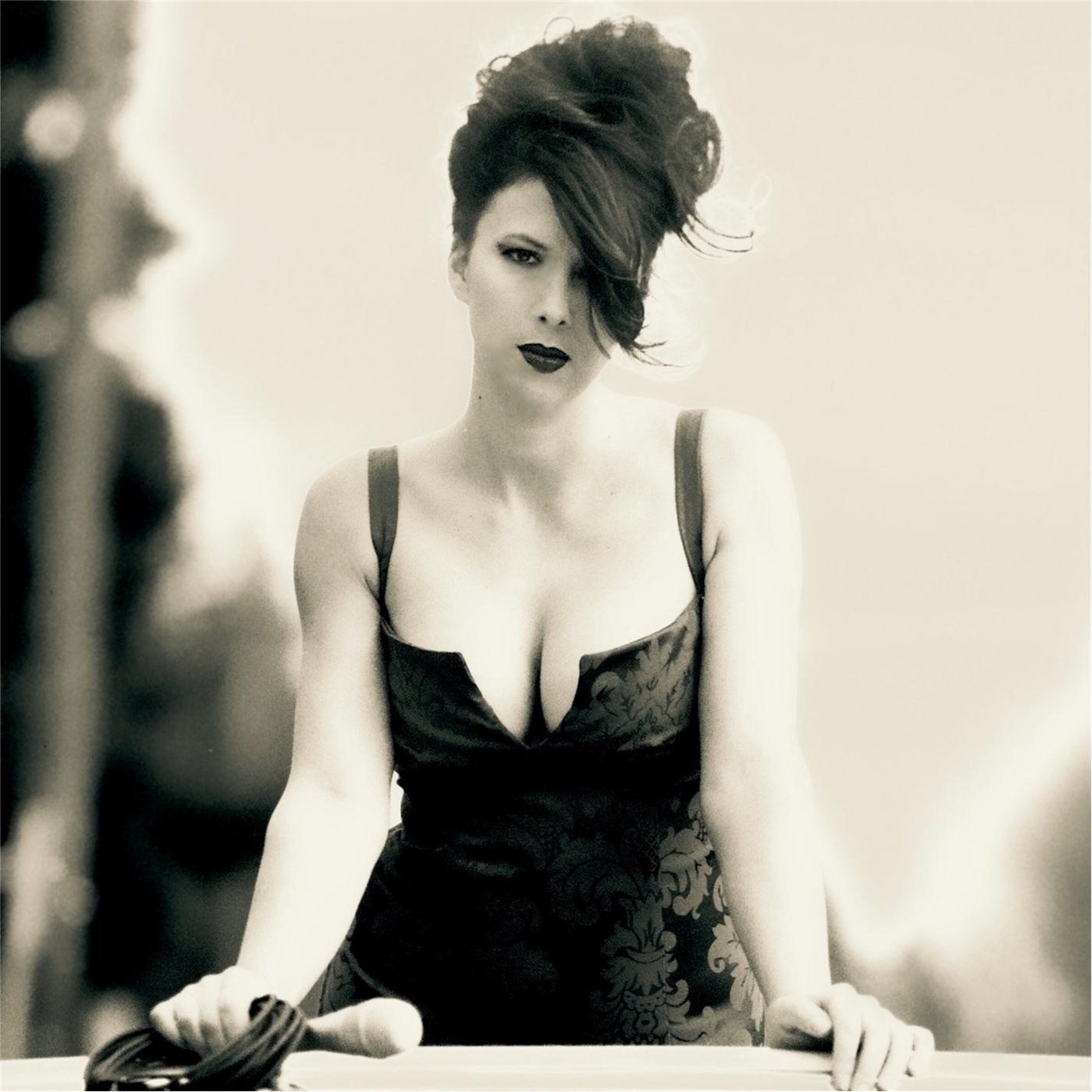 Black and white photograph of a woman with styled hair and dark lipstick, wearing a low-cut dress with floral patterns, leaning forward with her hands on a surface. She looks directly at the camera with an intense expression.