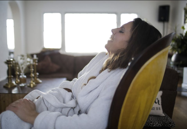 Tasha sitting in a chair, relaxing with her eyes closed and a content expression, wearing a white bathrobe in a well-lit living room.