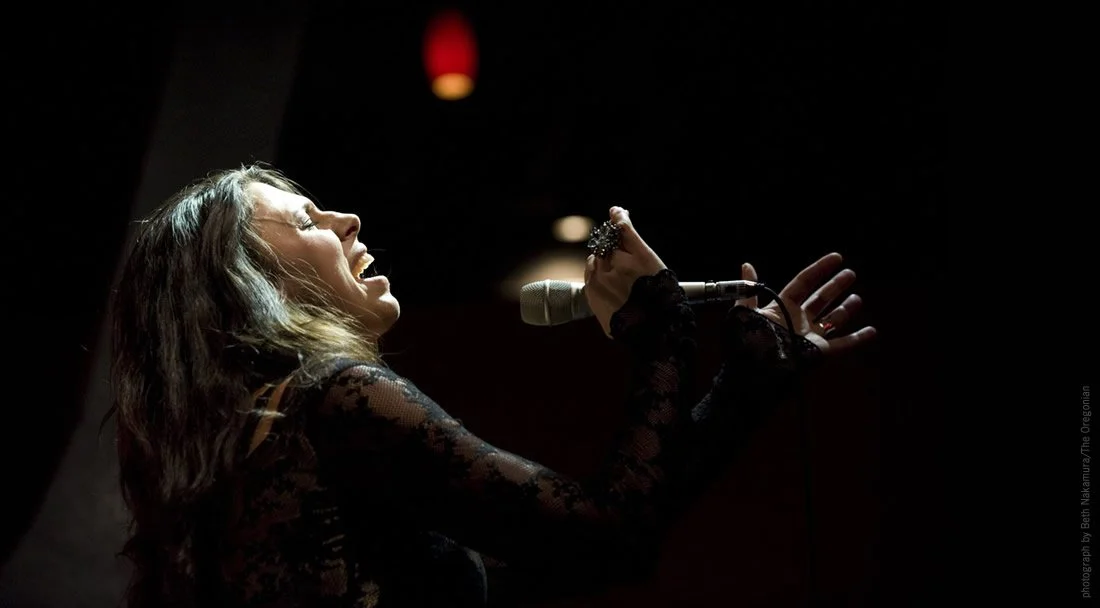 Tasha singing into a microphone in a dark setting, with a red light in the background.
