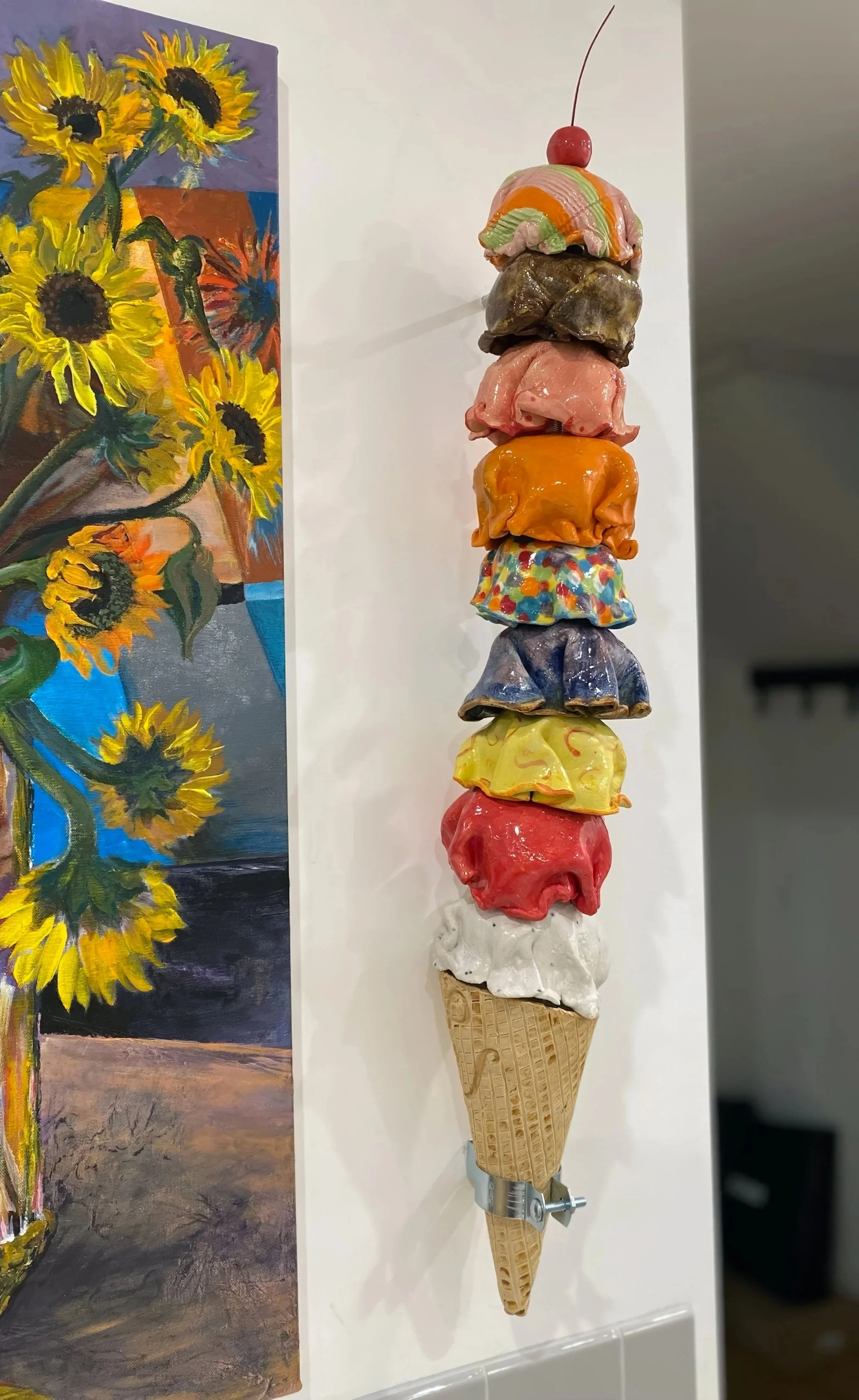 Colorful sculpture of a cone with multiple scoops of ice cream in different colors and textures, topped with a cherry and a cherry stem, displayed on a white wall next to a sunflower painting.