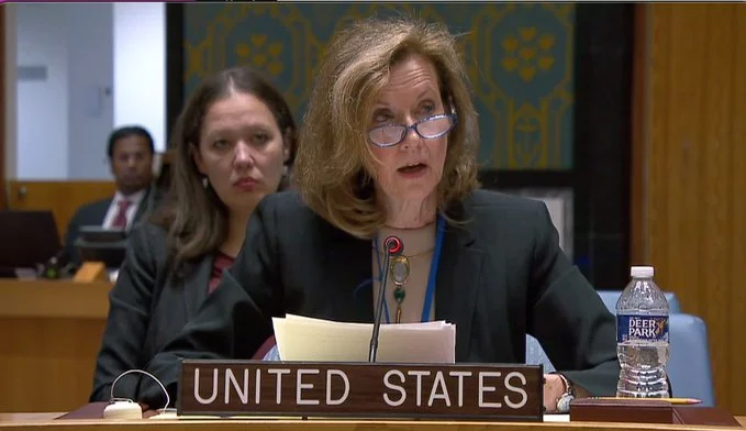 A woman speaking at a United Nations meeting with a 'United States' nameplate, wearing glasses, holding documents, and accompanied by two attendees in the background.