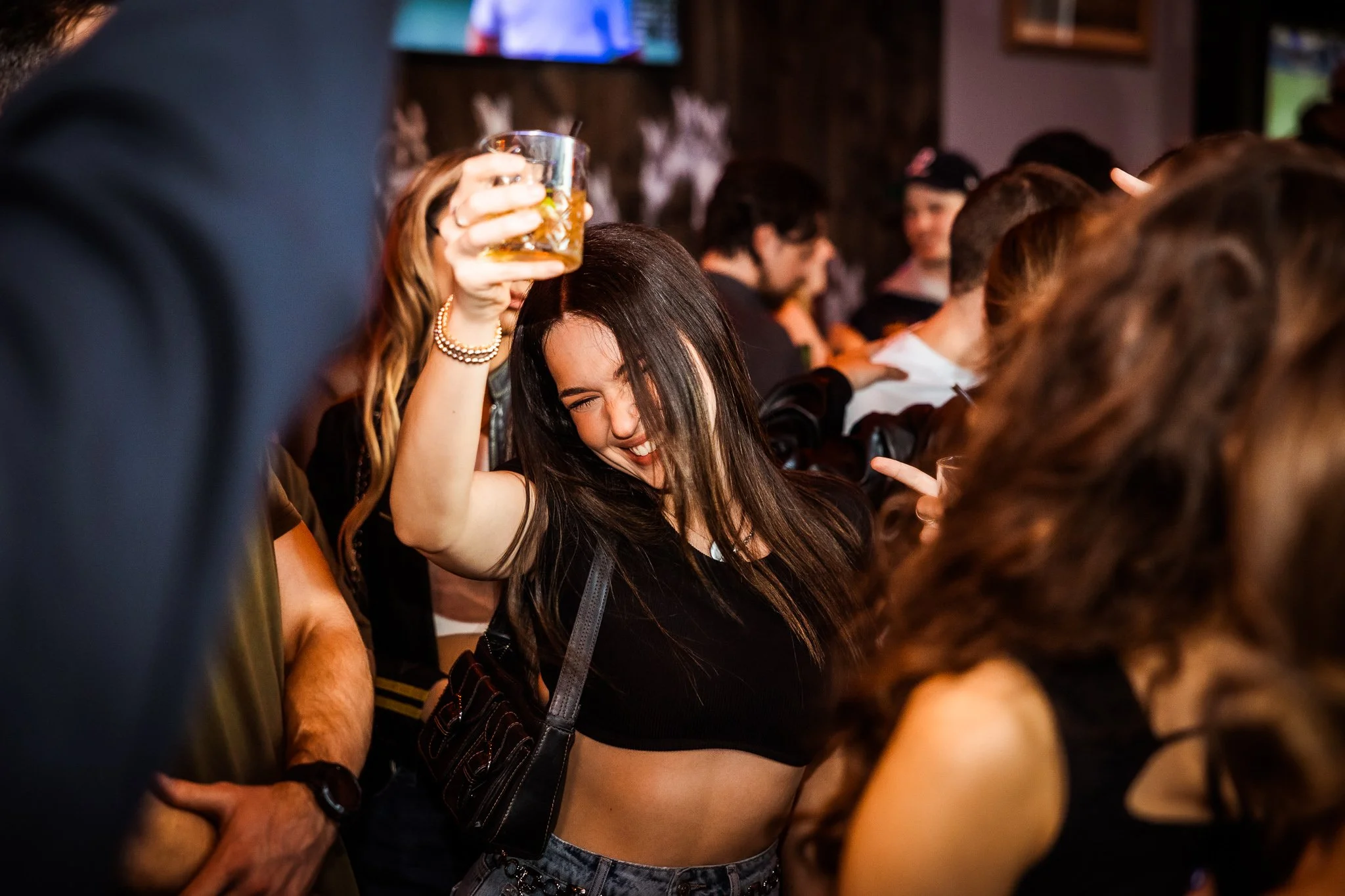 Woman dancing and holding up a drink at a party or bar with crowded background.